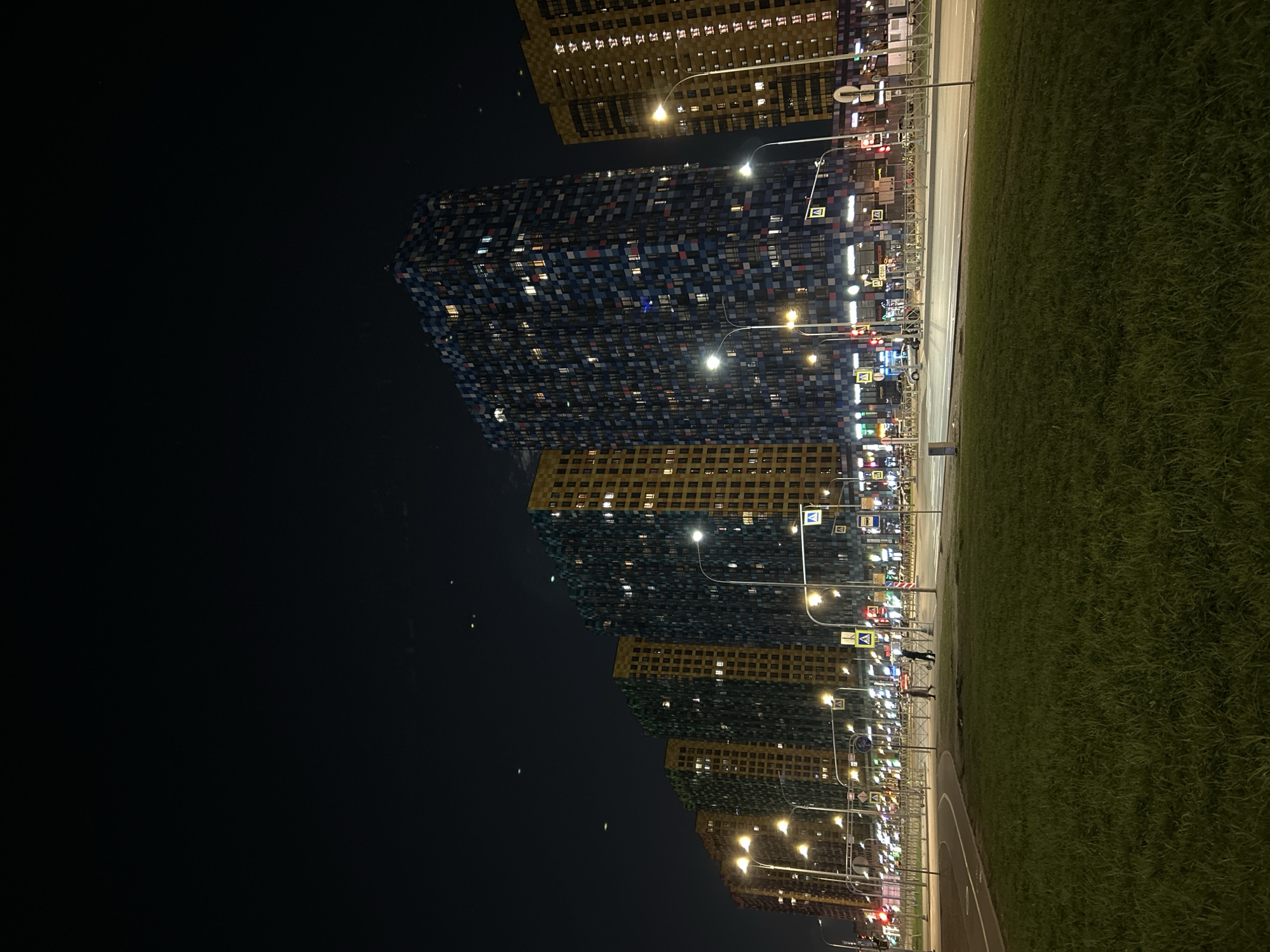 The image depicts a nighttime urban scene with a series of tall residential buildings, a multi-lane road, and a wide grassy strip. The perspective is from the grassy area looking towards the buildings.
The main subjects are several high-rise buildings illuminated by interior lights. Some buildings feature a distinctive checkerboard facade in shades of blue, grey, white, and red, while others have a more uniform yellow-gold exterior. Many windows are lit, suggesting active occupancy.
A well-lit road, possibly a multi-lane thoroughfare, runs parallel to the buildings. Numerous streetlights with bright, yellowish-white light illuminate the road, sidewalks, and the lower sections of the buildings. Traffic lights are visible in the distance, showing red and possibly other colors.
Separating the viewer's position from the road is a substantial grassy area, appearing as a neatly maintained lawn.
Several objects are present along the roadside: numerous streetlights on tall poles, a metal barrier or fence, and various road signs. Prominently visible are multiple blue square signs with a white triangle and a walking person symbol, indicating pedestrian crossings (common in Russia). Two indistinct human figures appear to be walking on the sidewalk near the road, illuminated by the streetlights.
The scene takes place at night, evidenced by the dark sky, the prominent illumination from artificial light sources (streetlights, building lights), and traffic lights. The sky is dark and clear, without visible clouds or precipitation. While specific text on shop signs or billboards is largely illegible due to distance and resolution, the distinct pedestrian crossing signs serve as a notable location cue consistent with the Saint Petersburg, Russia context. The overall atmosphere is that of a modern, bustling city after dark.