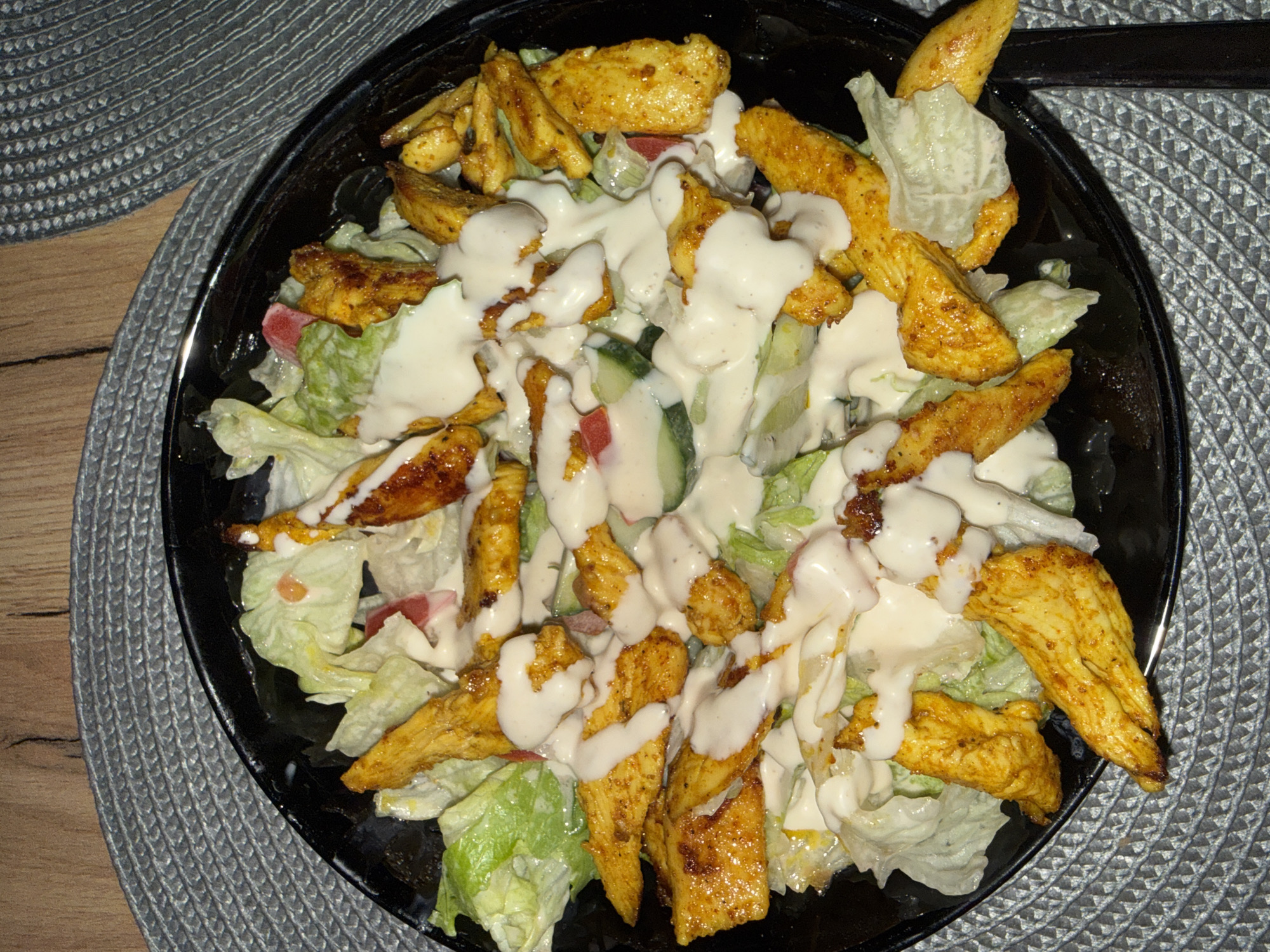 The image shows a close-up, overhead view of a salad in a black bowl. The salad contains lettuce, sliced cucumbers, some red pieces that appear to be tomato, and pieces of grilled chicken. A creamy white dressing is drizzled over the top. The bowl is placed on a grey, textured placemat, which is on a wooden table. The overall scene is casual and appears to be a meal setting. There is no indication of the specific location within the city of Varna, Bulgaria.