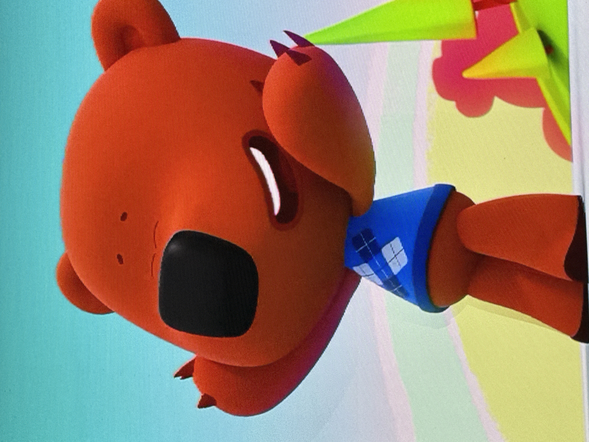 The image displays an animated, reddish-brown, bear-like character with a large black snout and notable black claws on its paws. The character is wearing a blue sleeveless sweater vest featuring a white argyle pattern. Its mouth is open in a crescent shape, conveying an expression that could suggest shouting, singing, or strong emotion. The character is positioned as if leaning into the frame from the right, with part of its body extending beyond the image edge.
The background is brightly colored and abstract, characteristic of a cartoon or digital animation setting. Visible background elements include sharp, pointed shapes in light green and yellow, alongside softer, rounded forms in light red and yellow. The lighting is bright and even, indicating either daytime or a well-illuminated indoor environment. The image captures an active moment within a colorful animated scene. There is no visible text or specific location cues beyond the general style of animation.