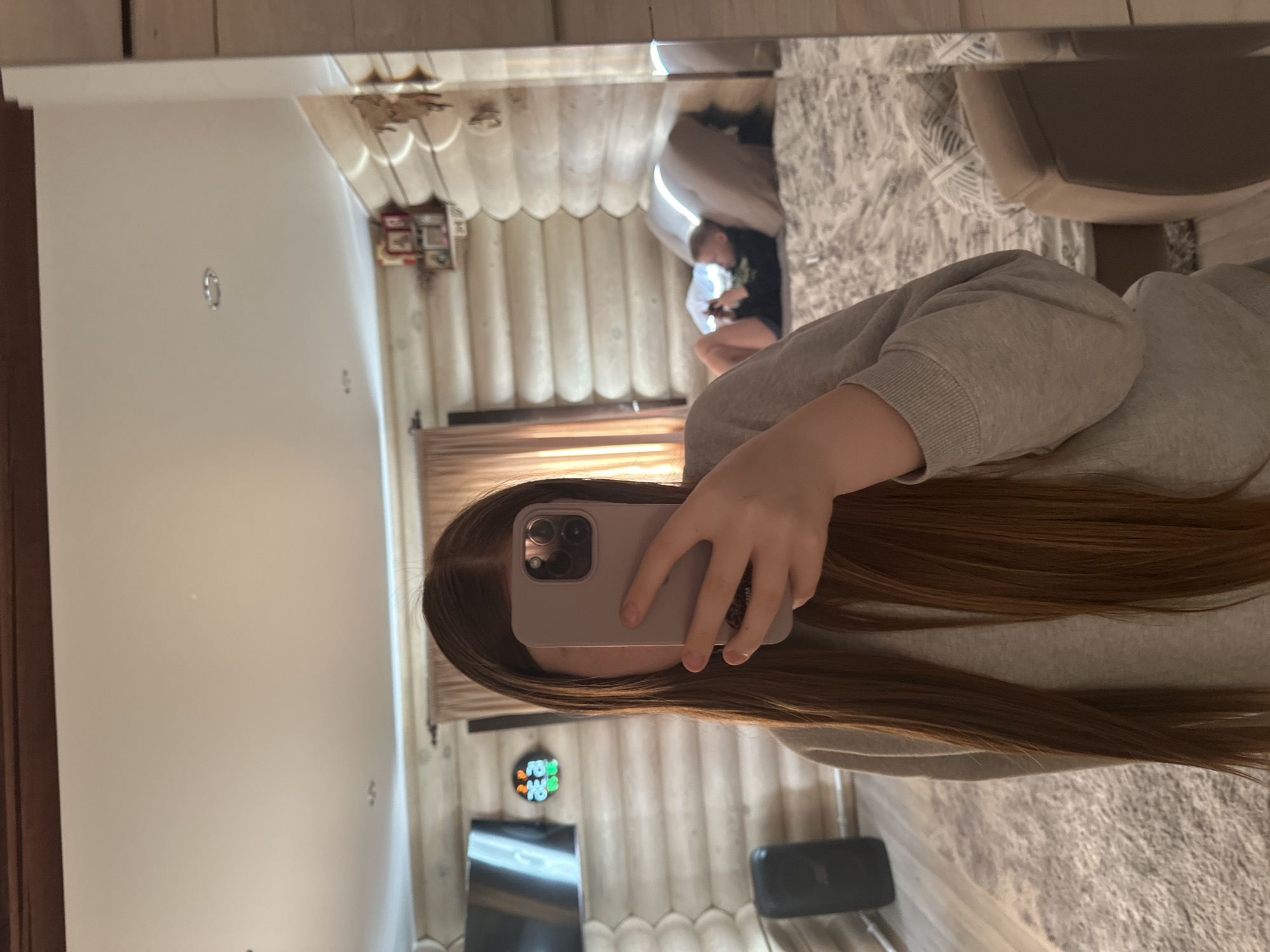 A selfie is taken in a mirror. The reflection shows a person with long brown hair holding a phone in front of their face, partially obscuring it. The phone is a light pink color with a triple-lens camera. The person is wearing a grey sweatshirt. In the background reflection, there is a glimpse of a living room with a beige couch, a white patterned rug, and a person seated on the couch using a phone. A TV and a clock are also visible on the wall. The location is Ольховка, Russia.