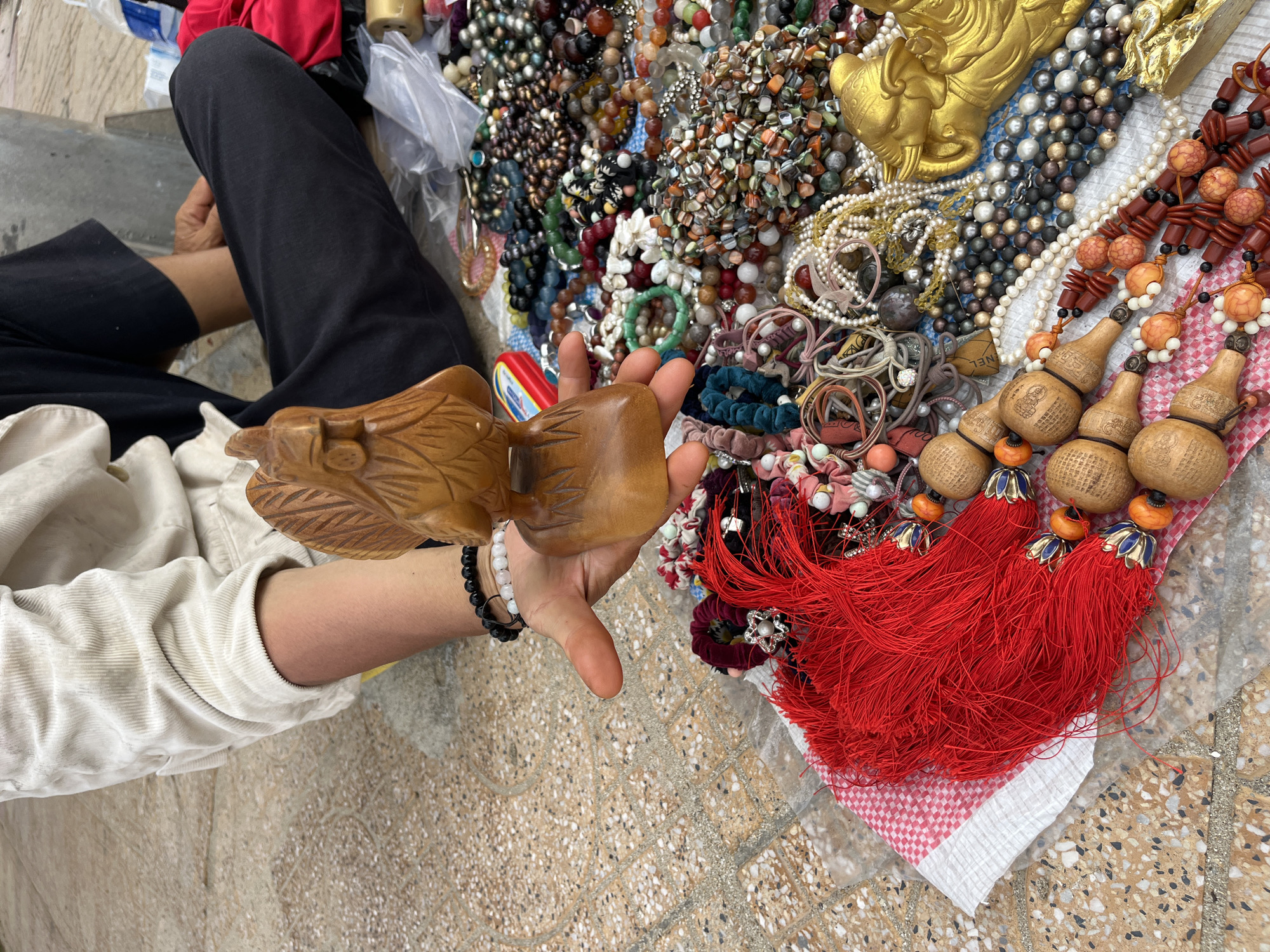 A person is sitting on a patterned tile surface, likely in a market or shop in the city of Bac Nha Trang Ward, Vietnam. The individual wears dark trousers and a light-colored, long-sleeved shirt. They are holding a carved wooden object in their right hand, displaying it among a collection of jewelry and other items laid out on a cloth. The items include strands of beads, necklaces, bracelets, and ornaments with red tassels. A golden elephant statue is visible. A variety of accessories like hair bands and other trinkets are scattered around, suggesting a market stall selling various items. The scene is densely packed with merchandise.