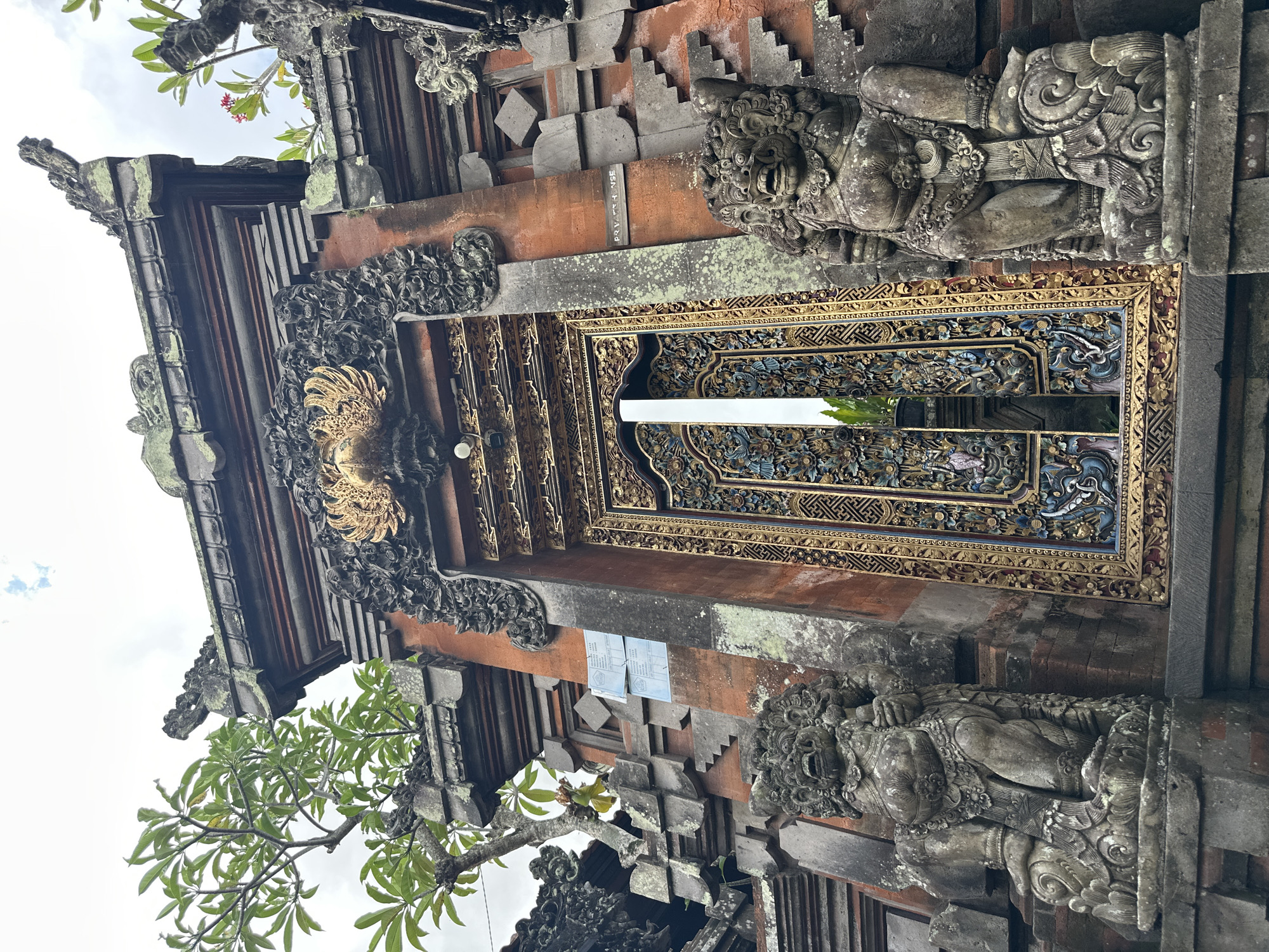 A low-angle, full shot captures an ornate entrance to a structure, likely a temple or traditional Balinese home, located in the Ubud District, Indonesia. The weathered, red-brick facade is adorned with intricate stone carvings, many of which are covered in moss, suggesting age and exposure to the elements. Two imposing stone statues of mythical guardians, with fierce expressions and bulky physiques, stand on either side of the entrance, each resting on a carved pedestal.
The central element is a richly decorated wooden door, framed by elaborate gold and dark carved panels. The door itself features a complex pattern of blue, gold, and other colored decorative elements, with a vertical reflective surface interrupting the center. Above the door, a golden eagle or phoenix-like emblem is prominently displayed, surrounded by dark, detailed carvings. The roofline of the structure is visible at the top, featuring more stone ornamentation and layered architectural elements. Green foliage from trees is visible encroaching on the structure from the left and right, with a few small red flowers blooming on the right. The sky above is overcast. A sign with text in a foreign language is affixed to the brickwork on the left side, and a smaller sign reading "PRIVATE HOUSE" is visible on the right side.