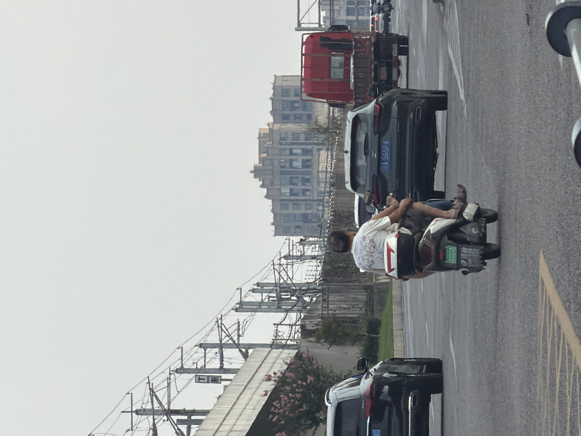 In the city of Jiangjin, China, on a street with multiple lanes of traffic, a black Mercedes-Benz SUV is stopped in the left lane. In the lane to its right, a person is riding a white and brown electric scooter, with another person seated behind them, their legs resting on the scooter's sides. The passenger appears to be a woman wearing shorts and sandals, and the driver is a man wearing a white t-shirt and shorts. Both are facing away from the camera. Further ahead in the right lane, a dark gray SUV with a license plate reading "A 5GSP4" is visible. Behind it, a large red flatbed truck is stopped, with a portion of a license plate reading "DE0107" visible. The road is paved with asphalt, and there are yellow lane markings. In the background, elevated train tracks with power lines stretch across the frame, supported by concrete pillars. A multi-story building with an unusual architectural style is situated beyond the train tracks, and a concrete wall with a fence separates the road from the elevated infrastructure. Lush green grass and some flowering pink bushes are visible along the side of the road. The sky is overcast.