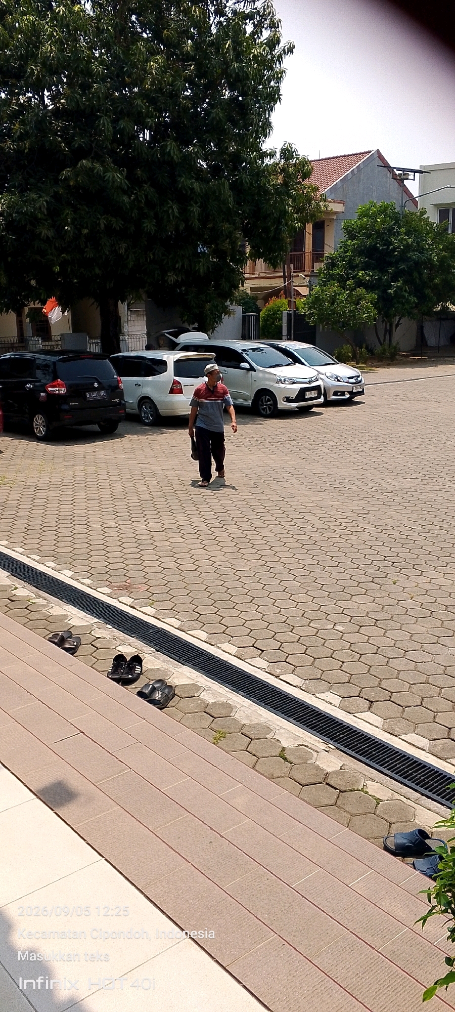 A man wearing a striped polo shirt, dark pants, and a cap walks across a hexagonal paved lot. He carries a dark bag in his right hand. Several cars, including a black SUV, a white minivan, and two white sedans, are parked behind him. A drain runs along the edge of the paved lot, separating it from a set of tan tiled stairs. Three pairs of sandals are placed on the ground near the stairs. The scene appears to be outdoors, under a clear sky, with trees and buildings visible in the background. The timestamp on the image indicates the date as 2026/09/05 at 12:25, and the location is identified as Kecamatan Cipondoh, Indonesia.