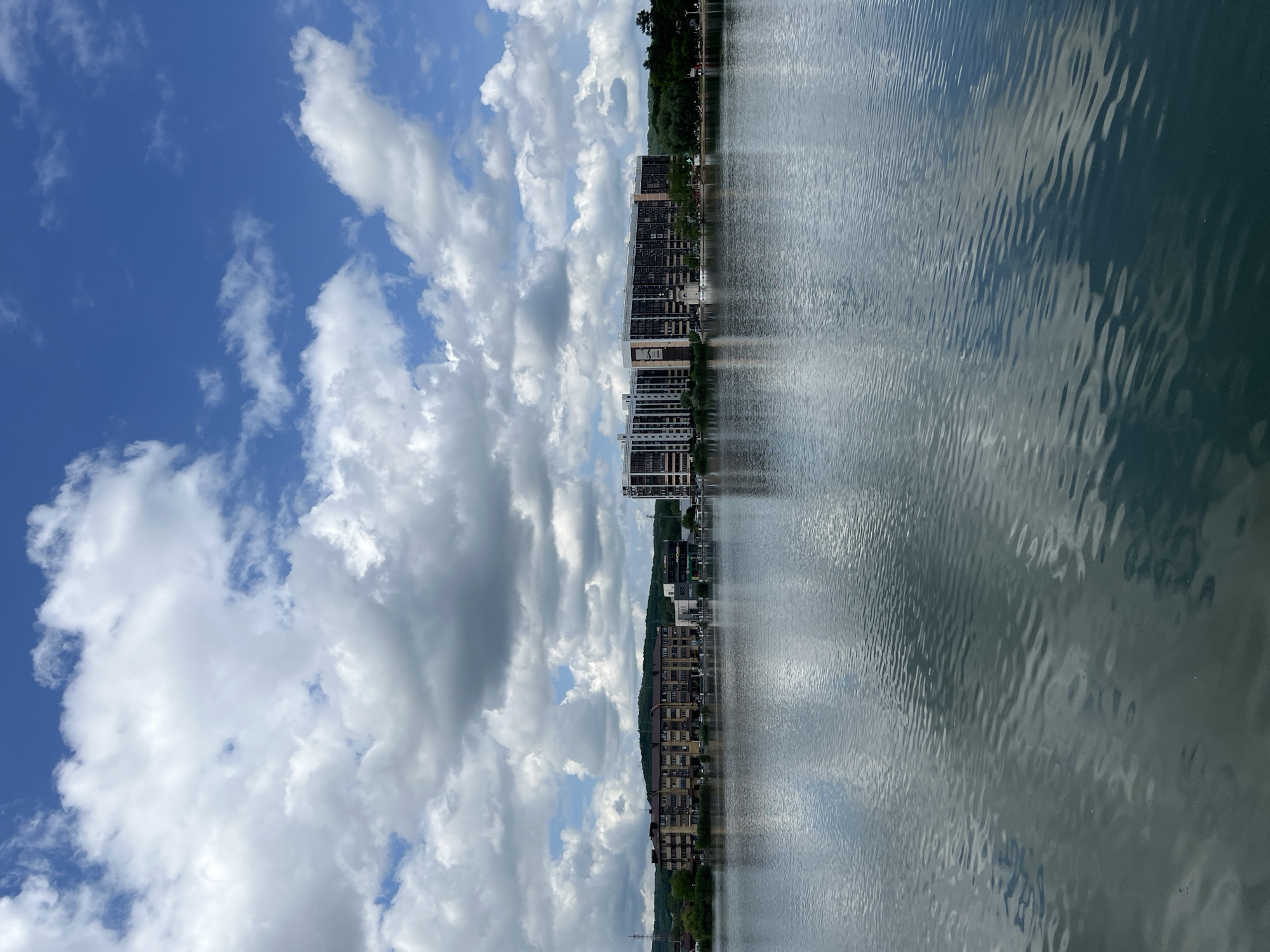 Here's a detailed description of the image:
The image depicts a waterfront scene, likely on a lake or river, in Goryachiy Klyuch, Russia. The water surface is rippled, reflecting the sky and the buildings on the opposite shore. The sky is partly cloudy with large, white cumulus clouds against a blue backdrop. Buildings of varying heights and architectural styles line the shore. Some are high-rise residential buildings with balconies, while others appear to be lower-rise structures. A strip of green trees is also present along the shoreline. The image appears to be oriented vertically.