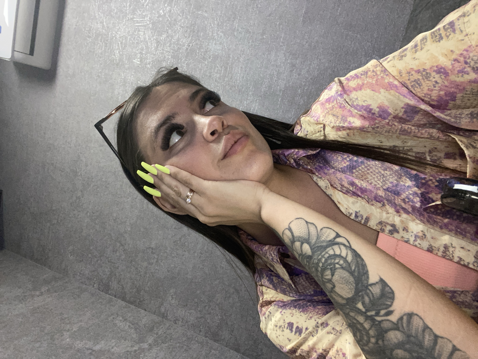 A young woman with long dark hair and a tattoo on her left arm is looking away from the camera, with her right hand covering part of her face. She is wearing a colorful, patterned shirt. Her fingernails are painted a bright, neon yellow. Glasses are perched on top of her head. The background appears to be a gray, textured wall, possibly in an interior space. There is a white object resembling an air conditioner or vent mounted on the wall above and to the right of the woman's head. The scene does not contain any identifiable landmarks or elements that would suggest a specific location within Makhachkala, Russia.