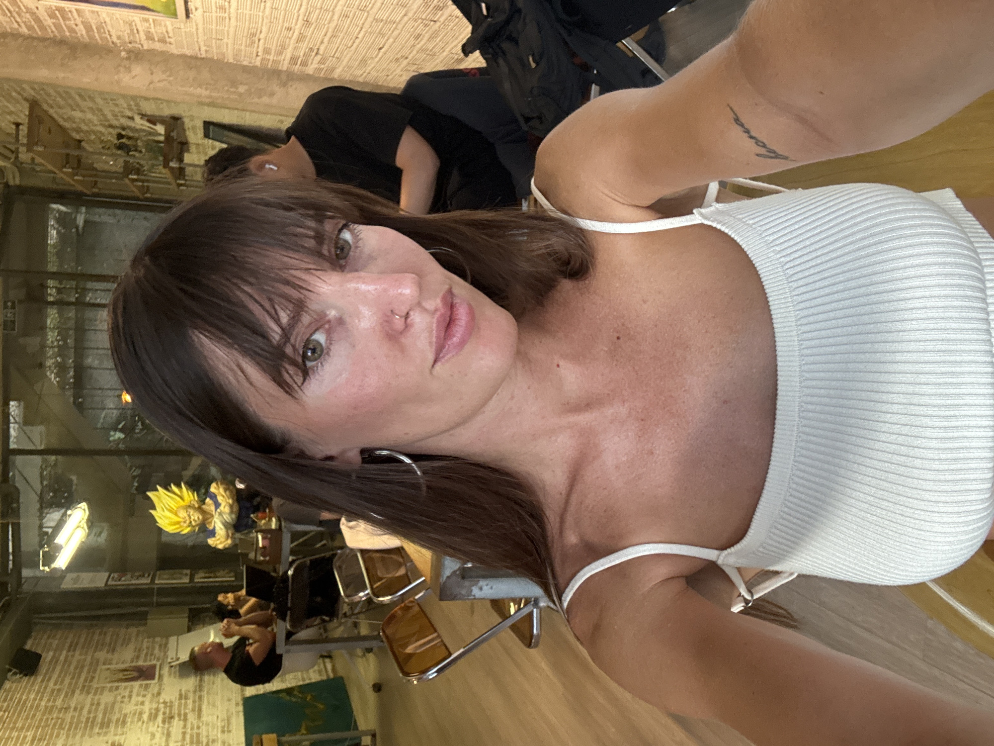 An indoor selfie captures a female subject, estimated 20-35 years old, with long dark brown hair and bangs, light skin tone, and green-brown eyes, in an establishment located in Da Nang, Vietnam. She wears a white ribbed crop top, large silver hoop earrings, and a small stud nose piercing on her right nostril. A faint cursive tattoo is visible on her upper left arm. The background reveals a dynamic space featuring a light-colored exposed brick wall on the left and a textured woven wall on the right. In the mid-ground, a vibrant Super Saiyan Vegeta figurine, characterized by spiky yellow hair and a blue outfit, is positioned on a table. To the left of the figurine, a male with short hair and a beard, wearing a black t-shirt, and a female with dark hair are seated at a table with an open laptop. A third, partially obscured individual in a black shirt is visible to the right. Overhead, a long, linear light fixture hangs, and a glass partition separates the depicted area from an adjacent space. The flooring is a light-toned wood, and industrial-style chairs with translucent amber backs are present. The environment suggests a modern cafe or co-working space.