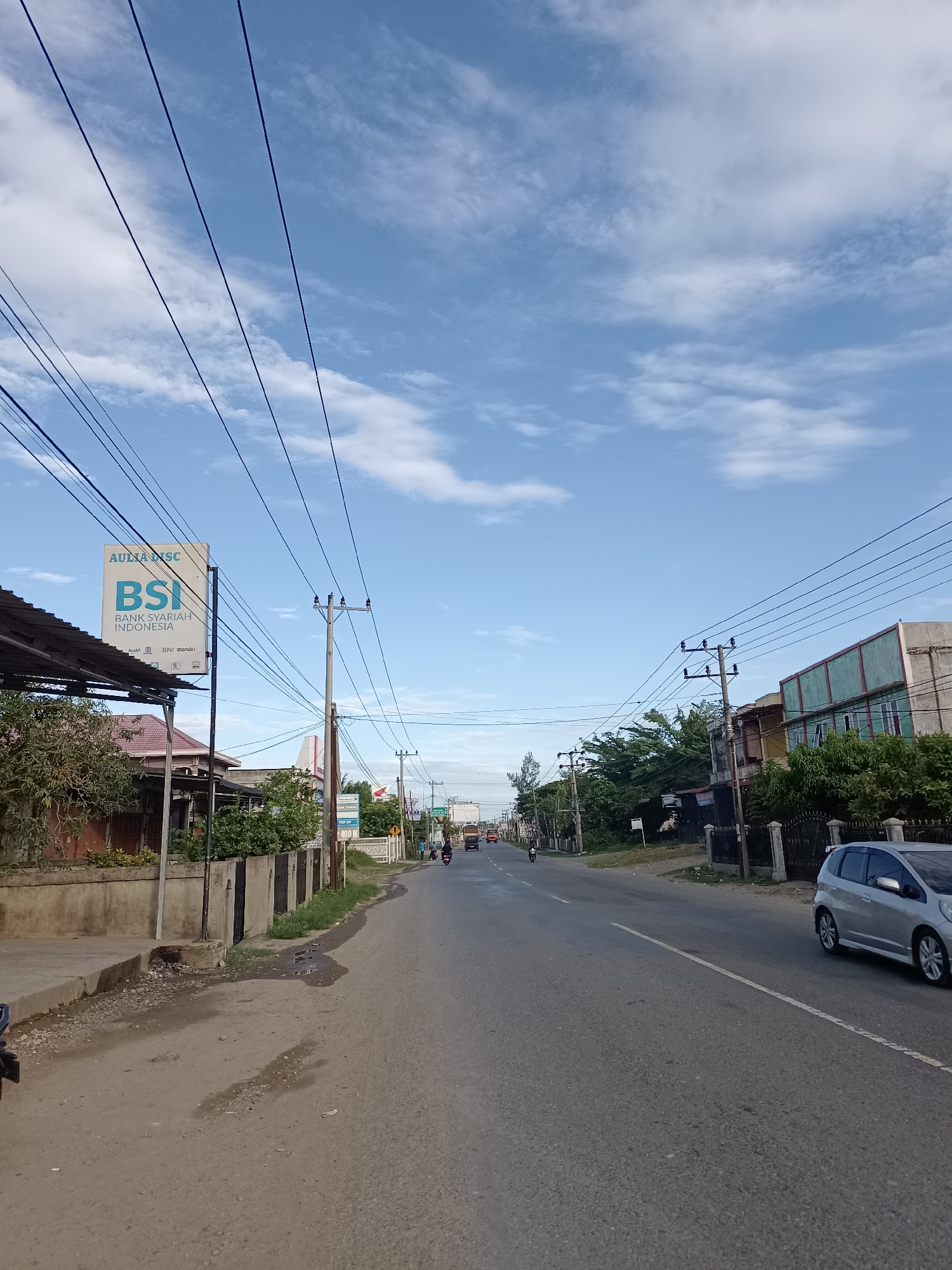 The image shows a street scene in Manyang Cut, Indonesia. A paved road runs through the center of the frame, with a sidewalk on the left and buildings and vegetation on both sides. Overhead power lines are visible across the sky. On the left side of the road, there is a sign for "AULIA MSC BSI BANK SYARIAH INDONESIA." Several vehicles are present, including a silver car on the right, a motorcycle, and a truck in the distance. The sky is blue with scattered clouds. The ground appears to be asphalt with some patches of dirt and puddles.