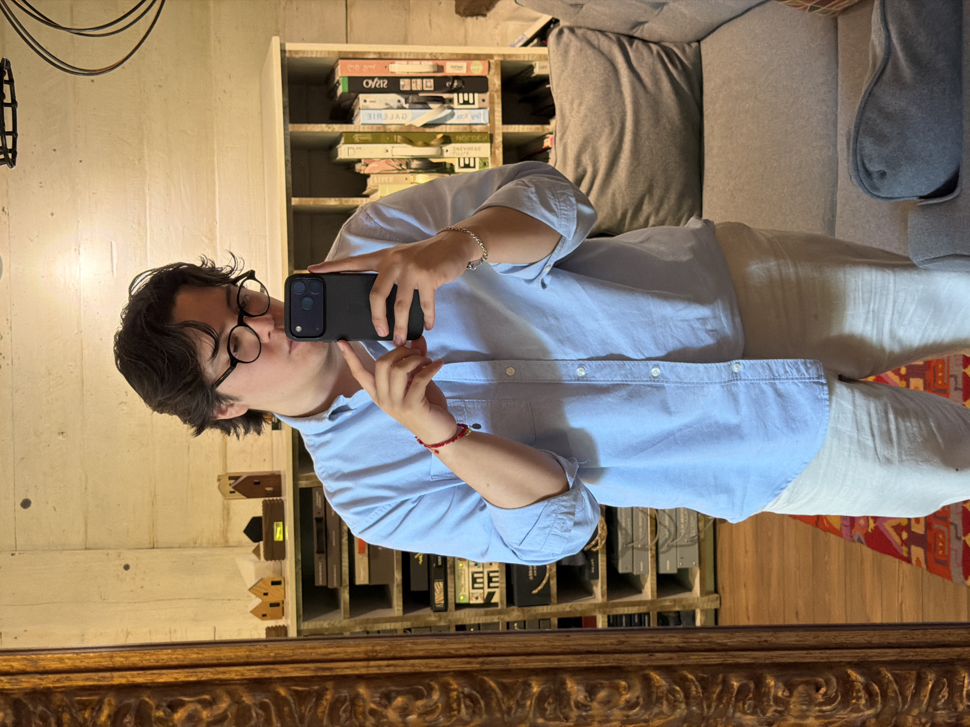 A person wearing glasses and a light blue button-down shirt is taking a selfie in a mirror. They are holding a black smartphone in front of their face with one hand. The person is standing indoors, next to a bookshelf filled with books and small decorative objects. A gray couch is partially visible in the background. The location appears to be in a home or apartment. The specific location in Puebla, Mexico, cannot be determined from the image alone.