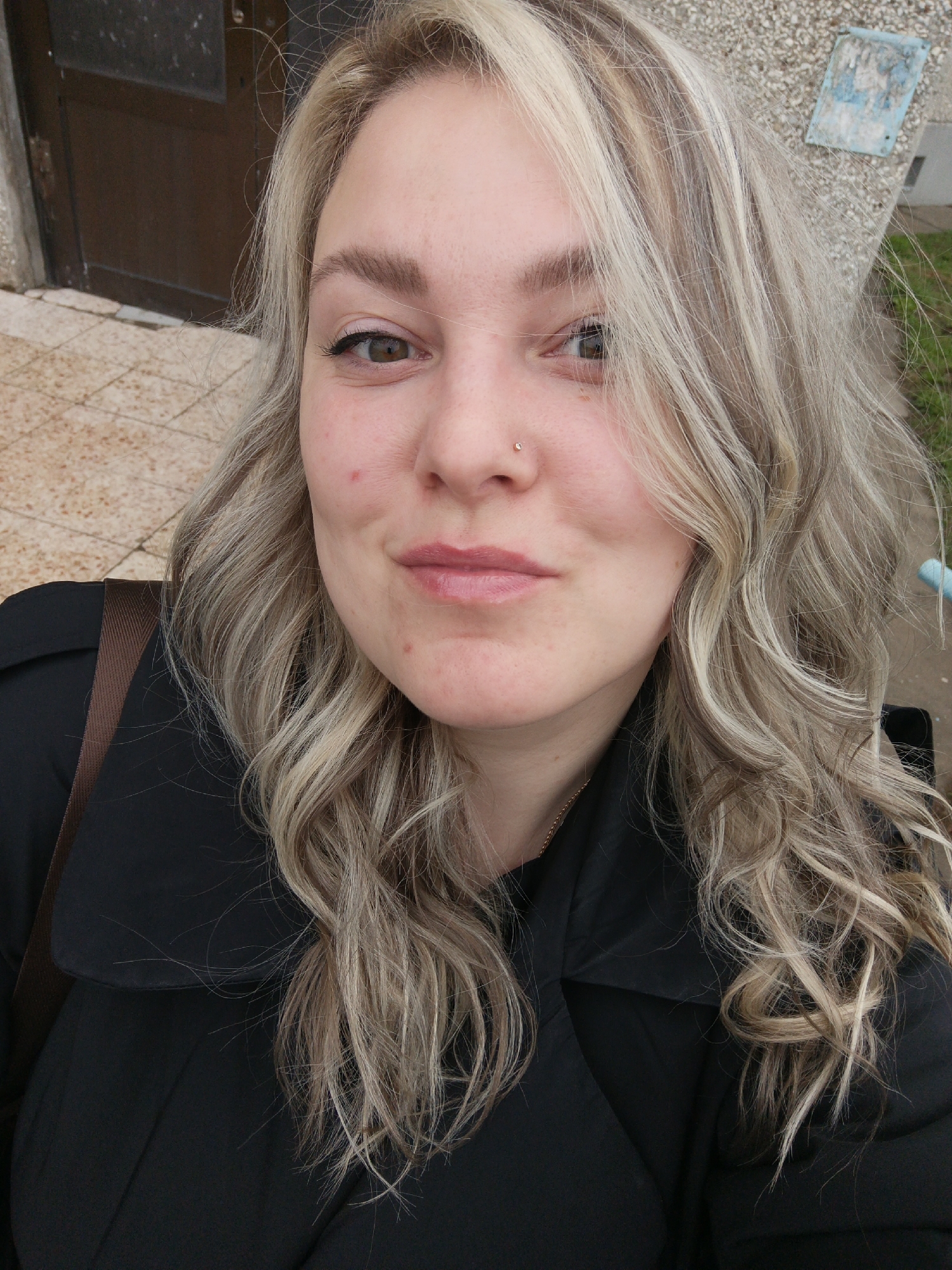 A woman with wavy, light blonde and brown hair, a nose piercing, and dark eyes is pictured in a medium close-up shot. She is wearing a black coat. The background includes a building entrance with a brown door and concrete steps. Part of a concrete wall or structure is visible on the right. The image appears to be taken outdoors during the day. There is no indication of being in the city of Марковский, Russia.