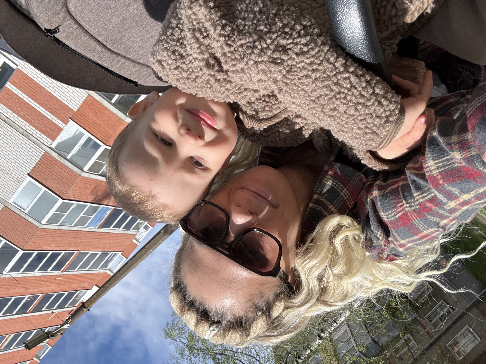 A selfie-style photograph captures a blonde woman wearing sunglasses and a red and gray plaid shirt, posing with a young child with blonde hair who is wearing a brown, fuzzy jacket. The woman’s hair is styled with braids at the front and loose waves. The child is nestled close to her, seemingly in an embrace.
In the background, an apartment building with a red brick facade is visible, along with a blue sky and a hint of green foliage. A portion of a stroller frame also appears in the image.
Given the information about the location, the scene is likely in Rusanovo, Russia, as specified. The image appears to have been taken outdoors on a sunny day, suggesting a casual moment captured while out for a stroll.
