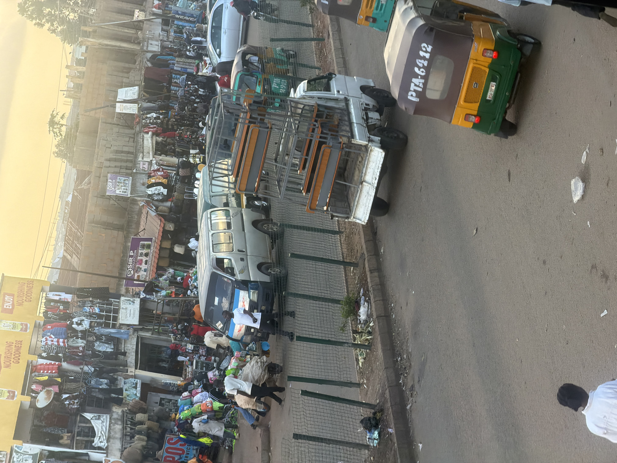This media file depicts a bustling street scene in Bukuru, Nigeria, likely in the late afternoon given the warm, low sunlight casting long shadows and illuminating the dust in the air. The image is vertically oriented, giving a sense of depth to the scene.
The foreground is dominated by a busy market area. Stalls overflow with various goods, primarily clothing and packaged food items, with racks of garments and piles of colorful merchandise visible. Several people are interacting within this space, some appearing to be vendors attending to customers, while others are browsing or in transit. Bags, sacks, and what look like rolled carpets are stacked around the stalls, suggesting active commerce.
The middle ground features a road with several vehicles. A white van, possibly a minibus, is parked near the market. Further down the road, a distinctive auto-rickshaw, often called a "keke," with the license plate "PTA-6412" is visible. Another open-air, multi-seater vehicle, likely a commercial transport, is also present, its seats arranged in rows.
The background shows more of the urban environment, including buildings under construction with visible brickwork, and other structures that form the backdrop to this lively market. The sky is a pale, hazy yellow, indicative of either sunrise or sunset, or possibly atmospheric conditions like dust or smoke.
Notable text includes "ENJOY NOURISHING GOODNESS" and "Malta" on signage in the market area, promoting food or drink. The text "ROS ERVICE" is also visible on what appears to be a business sign. The license plate "PTA-6412" on the auto-rickshaw is a clear identifying mark. The overall impression is one of a vibrant, everyday commercial hub in a Nigerian town, with activity and trade taking center stage under the warm light of the day.