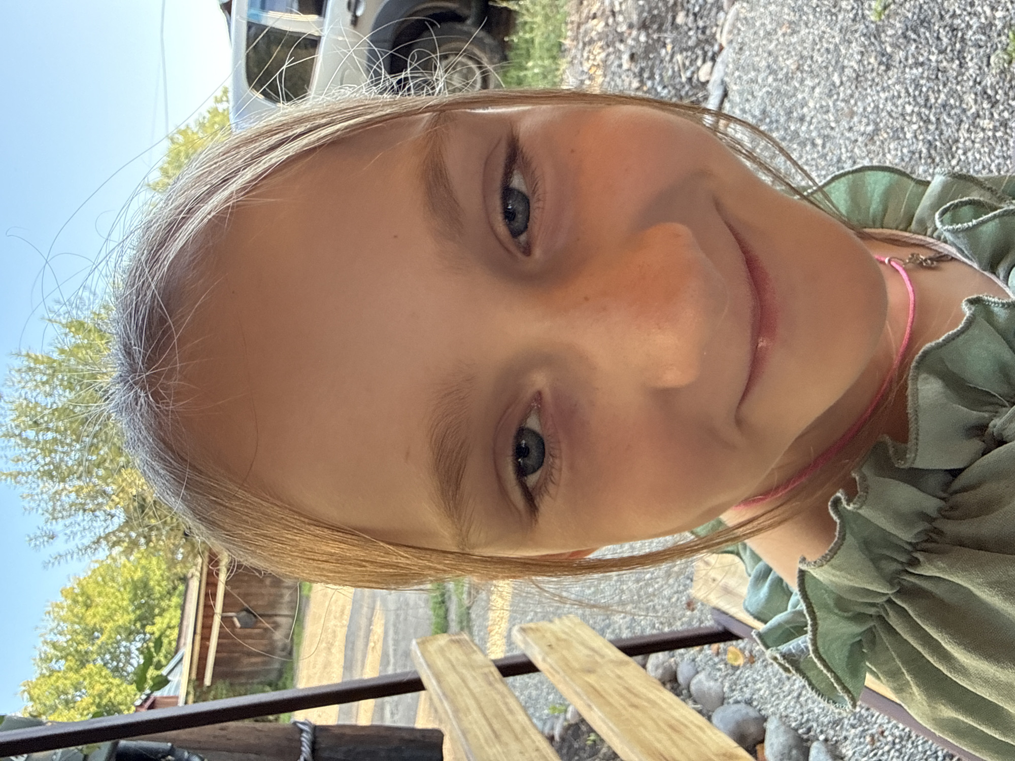A close-up, vertical, selfie-style photograph captures a young girl's face from the nose up, with her head tilted. She has fair skin with a scattering of freckles across her nose and cheeks. Her eyes are a light blue-gray, and she has thin, light brown eyebrows. Her blonde hair is slightly messy, with some strands falling across her forehead and face. She is wearing a green, ruffled top with a thin pink string visible around her neck. A faint smile plays on her lips, and her expression is one of mild amusement or curiosity.
The background is out of focus but suggests an outdoor setting. There are hints of green trees and foliage, a clear blue sky, and a gravelly ground. A portion of a silver vehicle is visible on the left side, and what appears to be a wooden fence or structure is on the far left. The lighting indicates it is daytime, likely sunny. The image appears to be taken outdoors in a residential or rural area, possibly in Krasnogorskoye, Russia, given the context. No text is visible in the image.