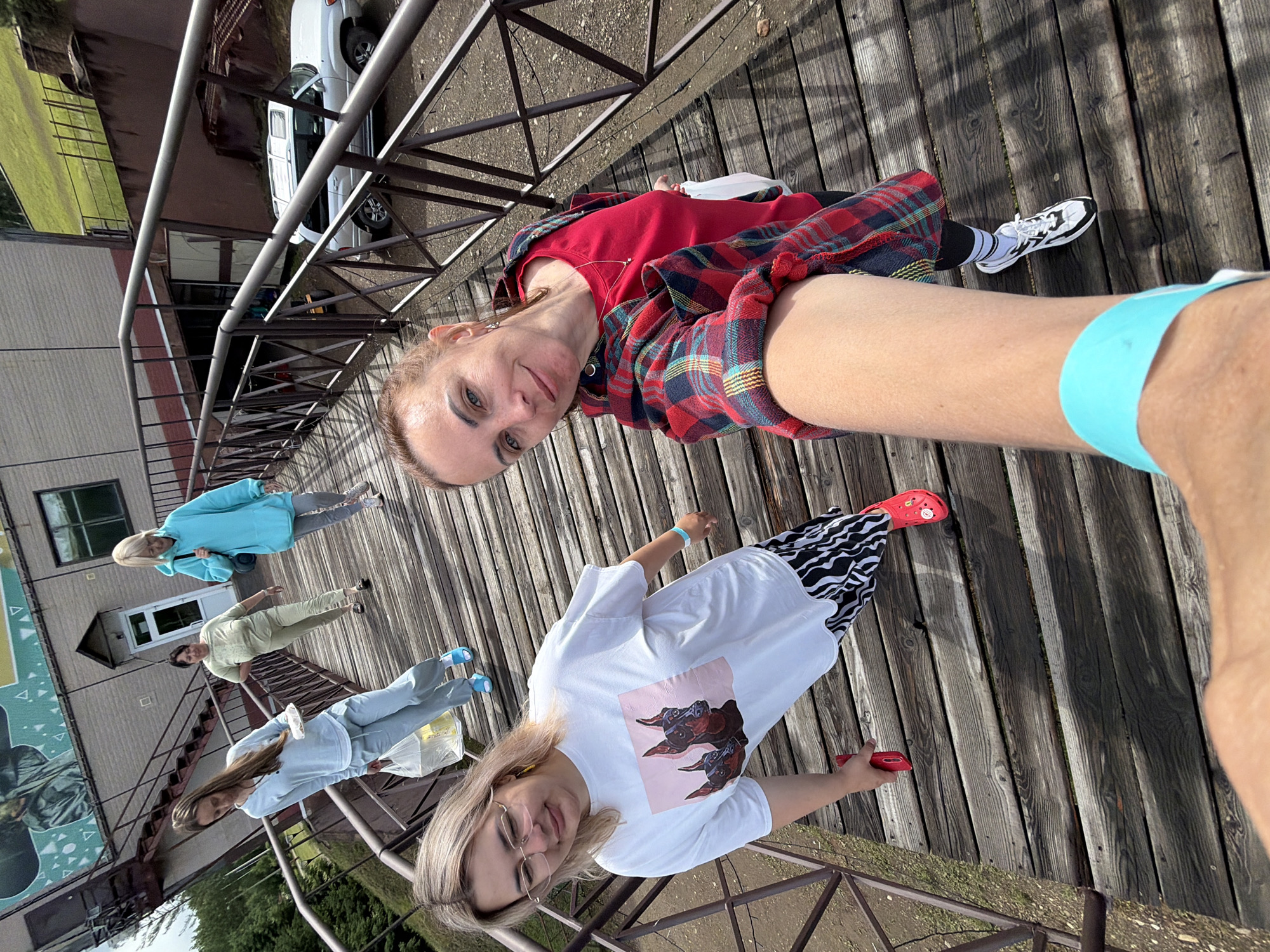 Four women are walking up a wooden walkway with metal railings. The walkway is outdoors, and in the background, there is a building with a mural on its side, a grassy hill, and a silver SUV. The women are dressed casually. The woman in the foreground on the right is taking a selfie. She is wearing a red shirt and a red and blue plaid shirt over it. The woman to her left is wearing a white t-shirt with a graphic of two dogs, black and white striped pants, and red shoes. Further up the walkway, a woman in a light blue hoodie and grey pants is walking, and behind her, a woman in a pale green tracksuit is walking. The woman in the front left is carrying a plastic bag with yellow contents and a white plate. The overall scene appears to be a casual outing in a semi-rural or recreational area. There is no identifiable information within the image to specifically place the location in Birobidzhan, Russia.