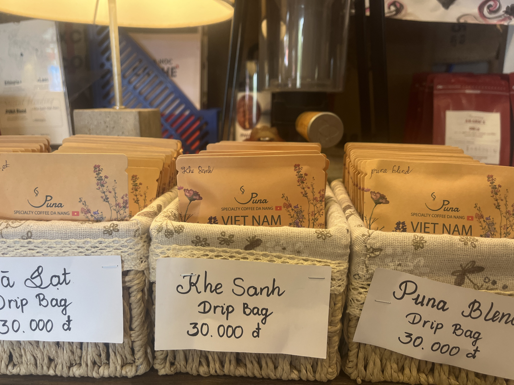 The image displays a close-up view of coffee drip bags arranged in woven baskets. Three distinct labels are visible, each indicating a different type of coffee and its price. The first label, on the left, reads "la Gat Drip Bag 30.000 đ". The middle label says "Khe Sanh Drip Bag 30.000 đ". The label on the right reads "Puna Blend Drip Bag 30.000 đ". The coffee bags themselves are small, individually packaged, and feature floral designs. The setting appears to be a retail environment, likely a coffee shop or market stall in Đà Nẵng, Vietnam, given the presence of Vietnamese currency and the explicit mention of "SPECIALTY COFFEE DA NANG" on one of the coffee bags. In the background, out-of-focus items like a lamp, a glass container, and other merchandise are visible. The lighting suggests an indoor setting.