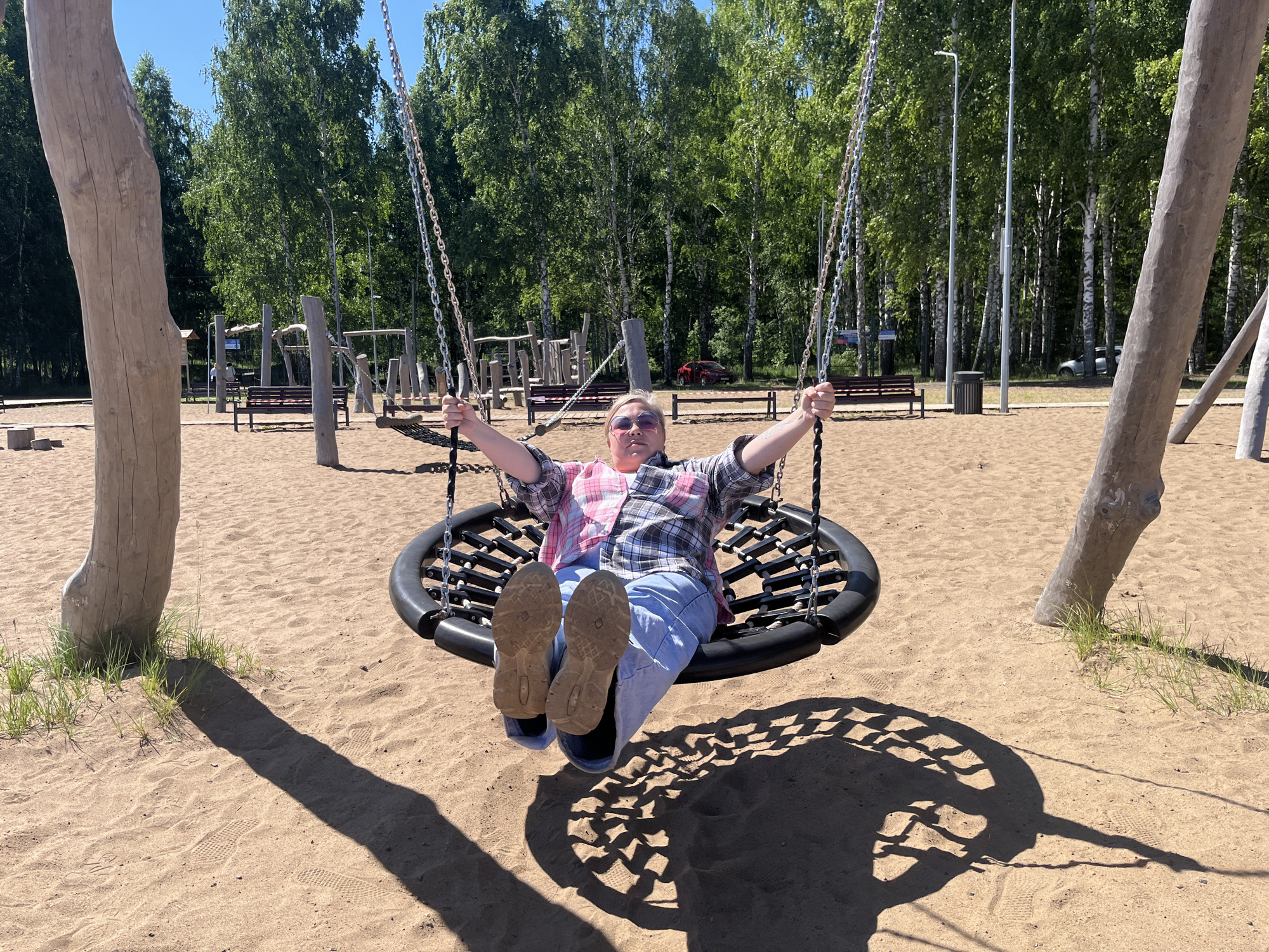 A person is sitting on a large, circular swing at a sandy playground. The swing is suspended by metal chains from a thick, light-colored log. The person is wearing a pink and plaid shirt, blue jeans, and light-colored shoes. Their arms are outstretched and their legs are bent. The playground appears to be located in a park with numerous birch trees and other deciduous trees in the background, suggesting a summer or early autumn setting. There are also some wooden structures and benches visible in the background, as well as streetlights and a trash can. The ground is covered in sand, and the shadow of the swing and the person is cast on the sand. The sky is blue and clear, indicating a sunny day. The image is taken from a medium distance, capturing the person on the swing and a portion of the surrounding playground and park environment. The location is specified as Galevo, Russia.