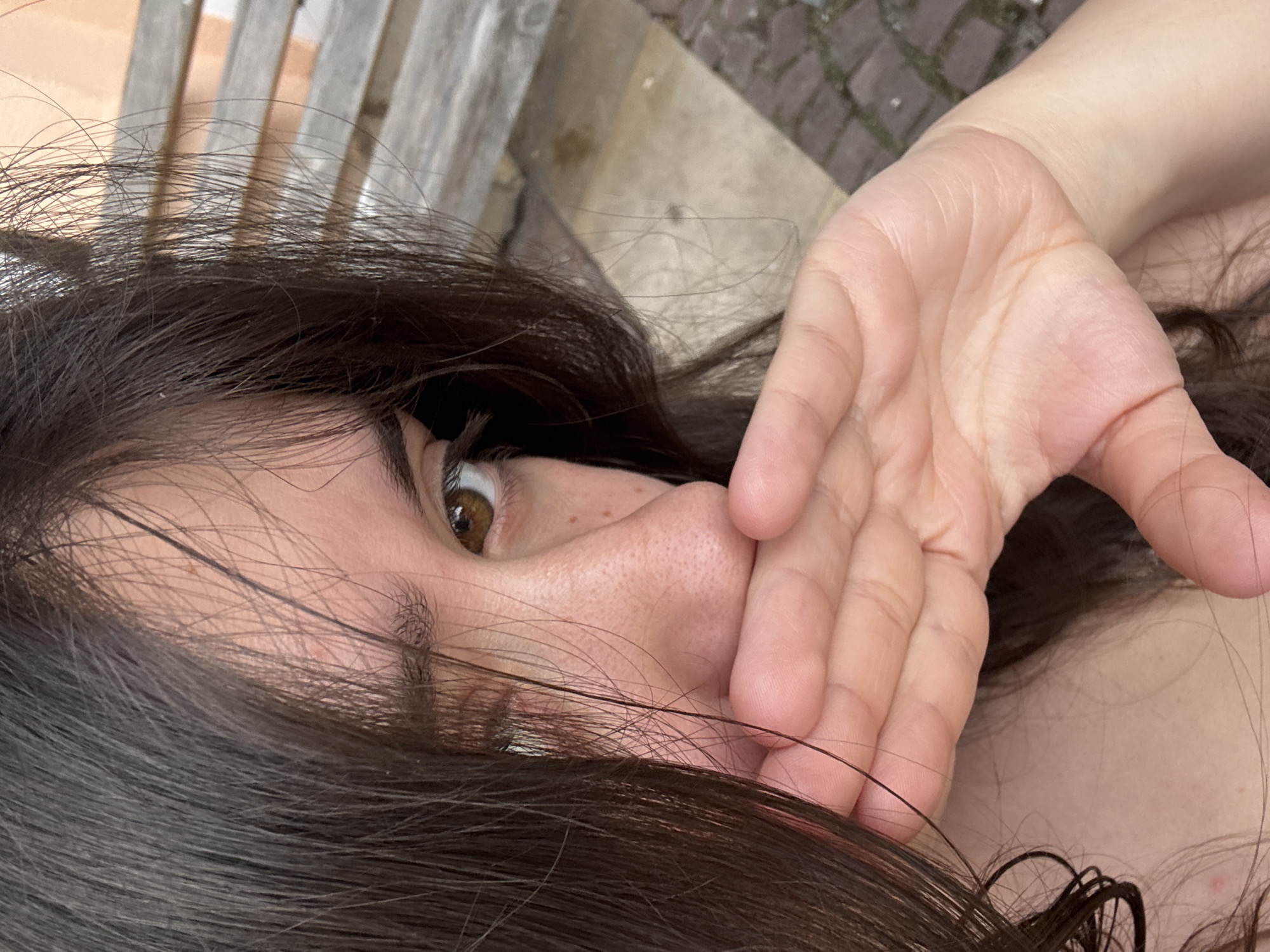 Close-up view of a person's face, primarily the left eye and surrounding facial features. The person has dark hair and light skin. A hand is positioned near the nose and mouth, partially obscuring the face. The background shows wooden slats of what appears to be a bench, and part of a stone-paved surface. There is no direct indication that the image was taken in Garmen, Bulgaria.