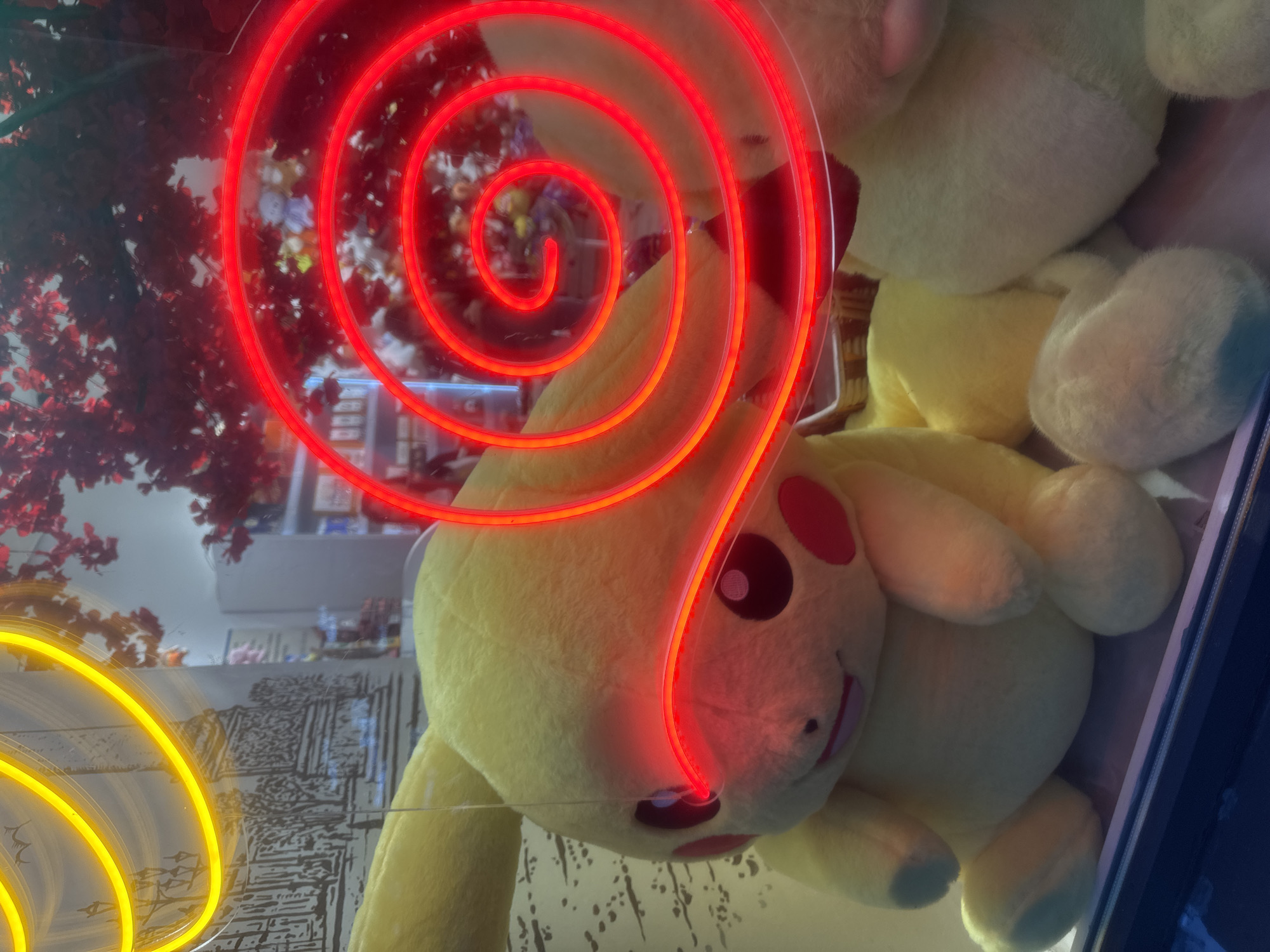 This image captures a large, yellow Pikachu plush toy positioned centrally behind a glass display, likely in a storefront or arcade. A prominent, bright red spiral neon light curves across the upper right and center, partially obscuring the background and passing directly over the Pikachu's left eye, causing it to appear red. In the upper left, a yellow circular neon light is visible.
Reflected in the glass in the upper left quadrant is a detailed, monochromatic architectural drawing or etching depicting a neoclassical building with a prominent colonnade and streetlights, strongly resembling the Kazan Cathedral in Saint Petersburg, Russia. Through the glass and around the red neon light, a retail interior is faintly visible in the background, showing shelves stocked with various merchandise, including what appears to be other plush toys, and a decorative artificial tree with red leaves. The scene is illuminated by the artificial light sources, creating a dynamic visual with the reflections.