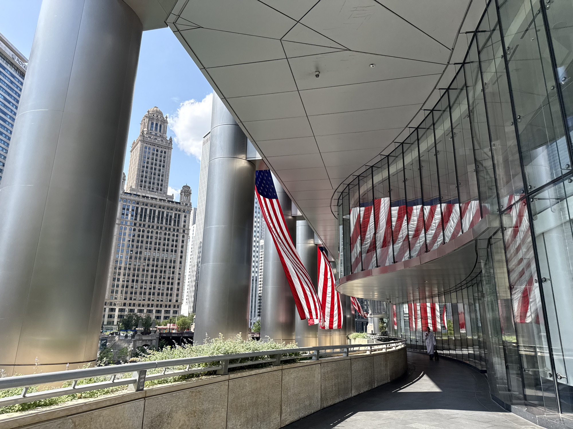 An outdoor walkway features a curved modern building with a glass facade displaying American flags as decoration. Large cylindrical columns support an overhang above the walkway. In the background, a distinct Art Deco skyscraper, possibly the Wrigley Building, stands among other city buildings under a bright sky with clouds. The walkway is bordered by a planter with greenery and a railing. One person, dressed in white, walks away from the camera down the curved path.