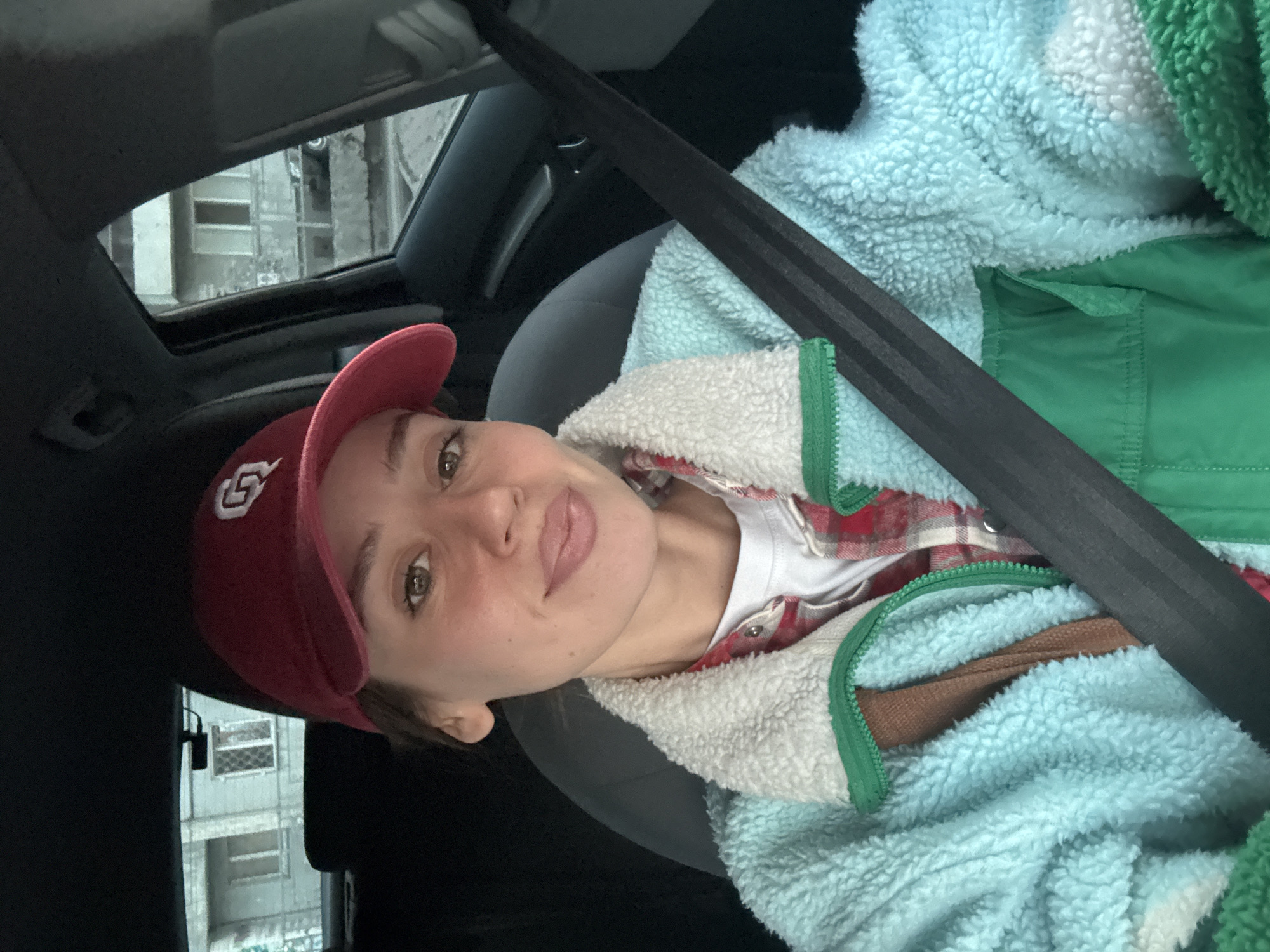 A person wearing a maroon baseball cap with white lettering is in the driver's seat of a car. The person is wearing a blue fleece jacket with a white sherpa lining and green trim, a red and white plaid shirt, and a white T-shirt. A black seatbelt is fastened across the person's chest. Through the car window, a building in Krasnoyarsk, Russia, is visible. The building appears to be grey and possibly residential, with windows and graffiti on the exterior. The scene suggests a candid snapshot taken from inside the car, likely during the daytime.