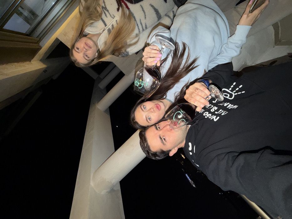 Three people pose for a selfie at night, each holding a wine glass. The person on the left has long blonde hair and wears a light-colored jacket. The person in the center has long brown hair and wears a light blue sweatshirt. The person on the right is wearing a black sweatshirt with white writing and a peace sign design. They appear to be outdoors on a balcony or patio.