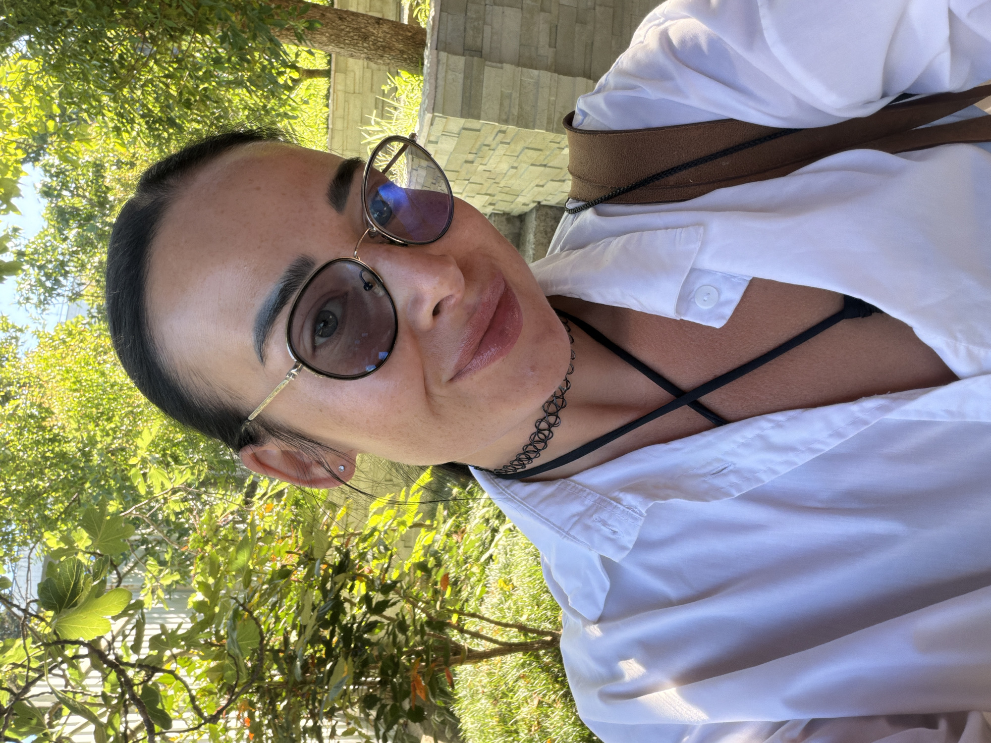 A woman with dark hair pulled back is taking a selfie outdoors. She is wearing round, gold-rimmed glasses and a white button-up shirt, unbuttoned to reveal a black strap bra and a black choker necklace. She is smiling, and her left shoulder is angled towards the camera. Behind her, green trees and foliage are visible, along with a stone wall and steps. The lighting suggests it is daytime, possibly late afternoon, with sunlight filtering through the leaves. The scene appears to be in a garden or park setting.