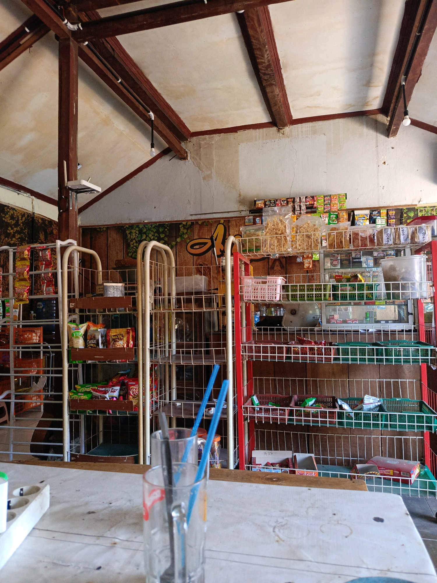 This is a low-angle, eye-level, medium shot of the interior of a small shop in Kebonagung, Indonesia. The shop appears to be a general store or warung, with shelves stocked with various food items and other merchandise. The walls are white, and the ceiling has exposed wooden beams and rafters, with some water stains visible. A single bare lightbulb hangs from the ceiling.
In the foreground, a white table is visible, with a white power strip with multiple outlets on the left side. A clear glass with two blue straws and some ice is placed centrally on the table, slightly out of focus.
Behind the table, metal shelving units are stacked with products. On the left, a tall, white metal shelf unit holds various packaged snacks and drinks. To its right, a red metal shelving unit is filled with more packaged goods, including what appear to be snacks, crackers, and possibly beverages. Some clear plastic containers and baskets are also present on these shelves.
The background of the shop shows more shelving units and a wall decorated with what looks like a mural or wallpaper depicting trees and foliage. The overall impression is of a simple, functional retail space. No people are visible in the image.
