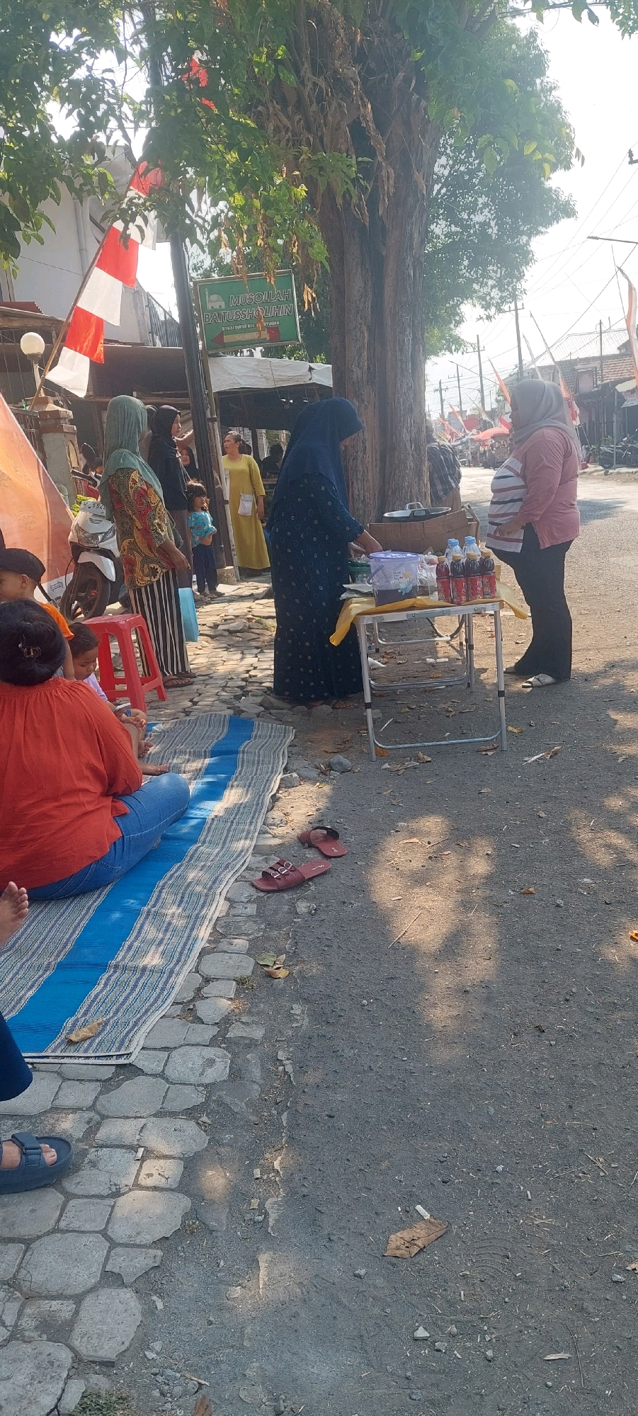 The image depicts a street scene in Jogoyudan, Indonesia. Several individuals are present, including women wearing hijabs and a child. One woman in a blue hijab and dress is interacting with a small table set up outdoors, appearing to be a vendor. On the table are several bottles of drinks and some packaged items. Another woman, pregnant and wearing a pink and white striped shirt and dark pants, stands near the table. In the background, a motorcycle is parked next to a fence adorned with Indonesian flags. The ground is paved with interlocking stones in some areas and dirt/gravel in others. The environment suggests a warm, sunny day. A sign with text in Indonesian is visible on a pole behind some of the individuals. There are also some red plastic chairs and a blue and white striped mat laid on the ground, where people might be sitting.