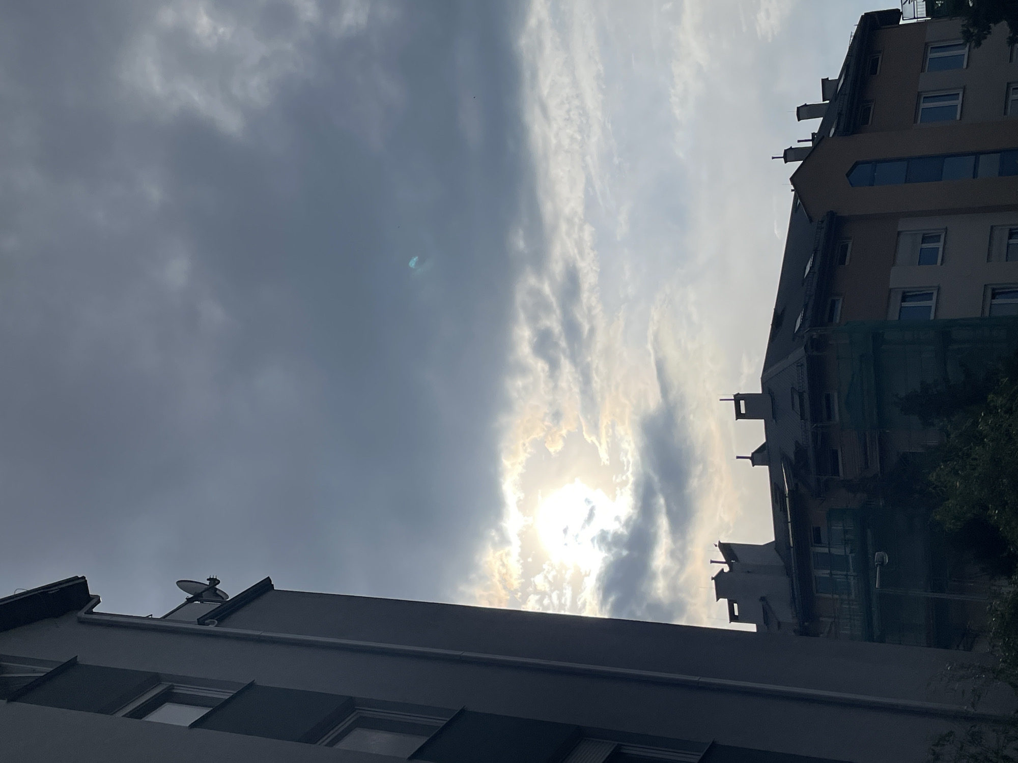 The image shows a low-angle shot of buildings against a cloudy sky. A building on the left has a flat roof with windows and a satellite dish. The sky is predominantly gray with a patch of sunlight breaking through the clouds in the center. On the right, a taller building with multiple stories and visible windows is partially obscured by trees. No people are visible. The location appears to be an urban environment.