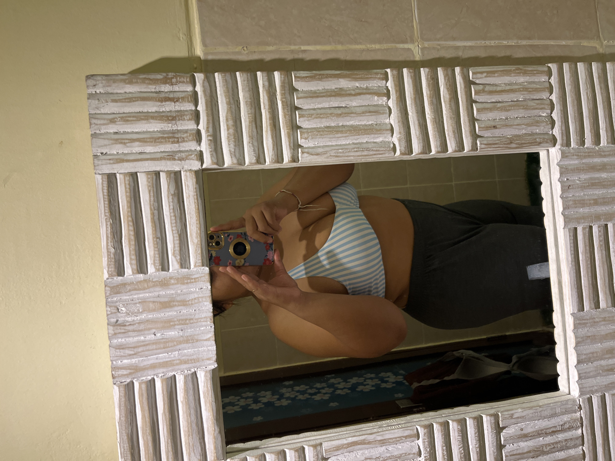 A person is taking a mirror selfie in a bathroom. They are wearing a blue and white striped bikini top and dark gray sweatpants. The mirror has a distressed white wooden frame with a textured, ridged design. The bathroom walls appear to be tiled in a beige color. In the reflection, a wall with a floral pattern can be seen behind the person. The overall setting is a personal bathroom.