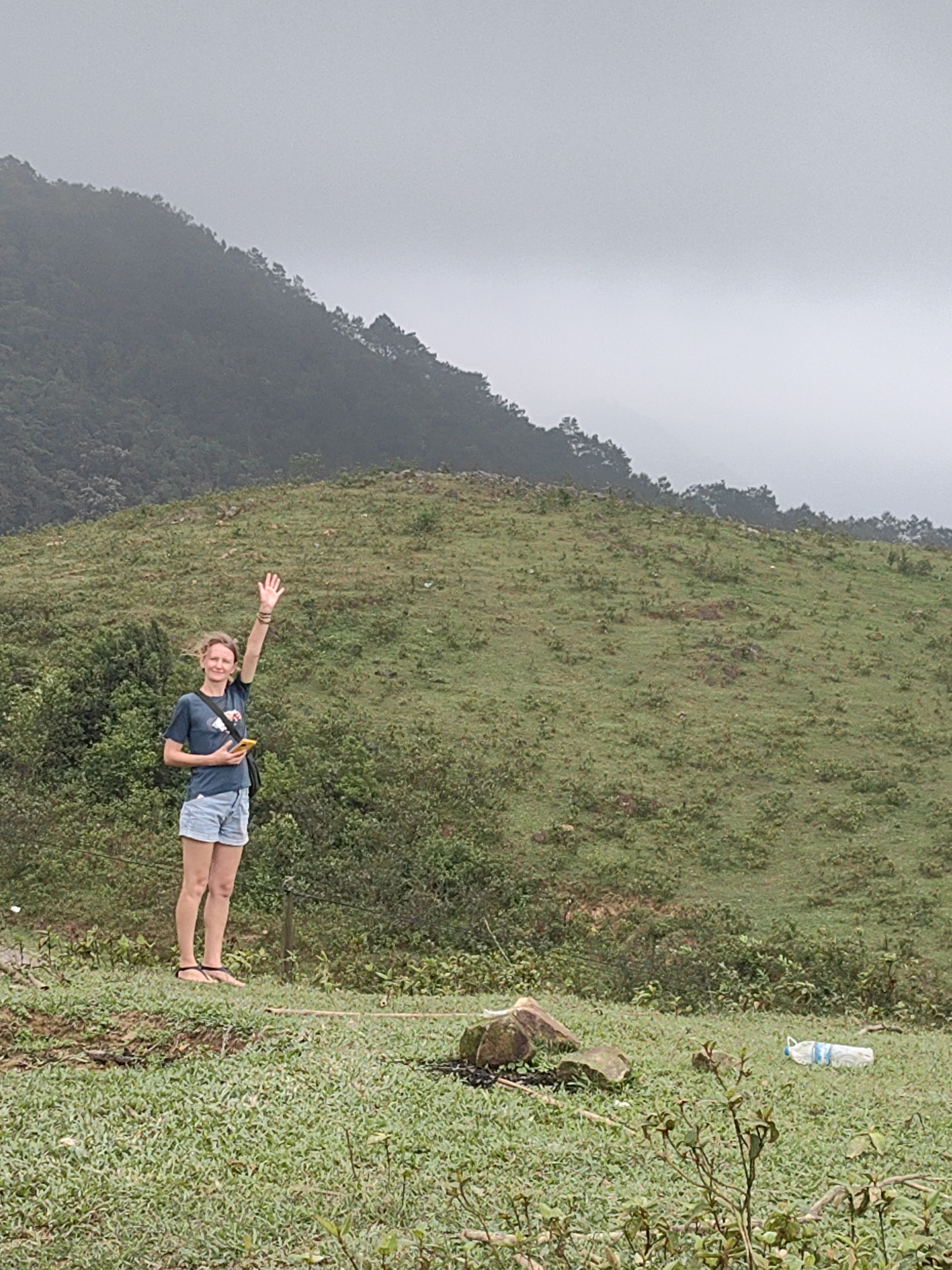 The image displays a light-skinned woman standing on a grassy hillside, waving with her right hand. She has medium-length blonde hair, is smiling, and appears to be in her 30s. She is dressed casually in a dark blue t-shirt with a graphic design, light blue denim shorts, and dark sandals. A dark shoulder bag hangs across her body, and she holds a yellow mobile phone in her left hand.
The scene is set in a natural, hilly landscape, specifically Thong Nhat Commune, Vietnam. The background features mountains covered in dense, dark green trees and foliage. The sky is overcast and grey, and a light mist or haze partially obscures the mountain peaks, suggesting a cloudy or humid day. The ground in the foreground and midground is covered in varying shades of green grass and small shrubs. In the lower right portion of the image, a discarded light blue plastic water bottle lies on its side near some rocks and patches of bare earth. The overall impression is one of outdoor activity in a scenic, mountainous region under diffuse natural light. There is no visible text in the image.