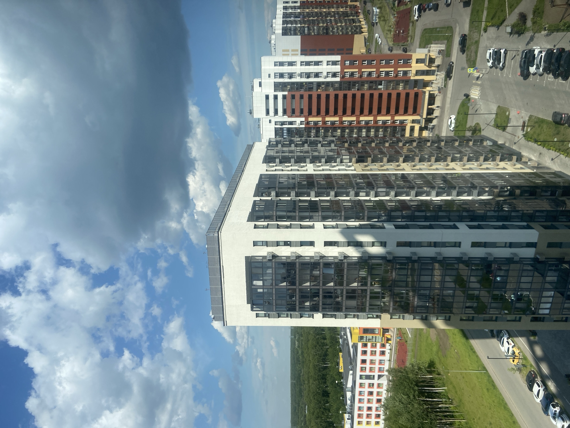An aerial view captures a modern residential complex in Kazan, Russia, under a sky with scattered white and dark gray clouds. The complex features a cluster of tall, contemporary apartment buildings with white, beige, and reddish-brown facades. The buildings have numerous windows, many of which are reflective, showing the sky and surrounding structures. Some buildings are segmented by color, with prominent vertical bands of red and beige on some facades. One taller building in the foreground is primarily white with a large section of dark-tinted glass balconies. Adjacent to the apartment buildings is a lower structure with a colorful facade, featuring red, yellow, and white sections, and a playground with red and yellow equipment. A road runs alongside the buildings, with parked cars, including white sedans, black SUVs, and a small yellow car. The area is bordered by a dense green forest to the left and a neatly mown grassy area with some trees. The scene is characterized by urban development set against a natural backdrop.