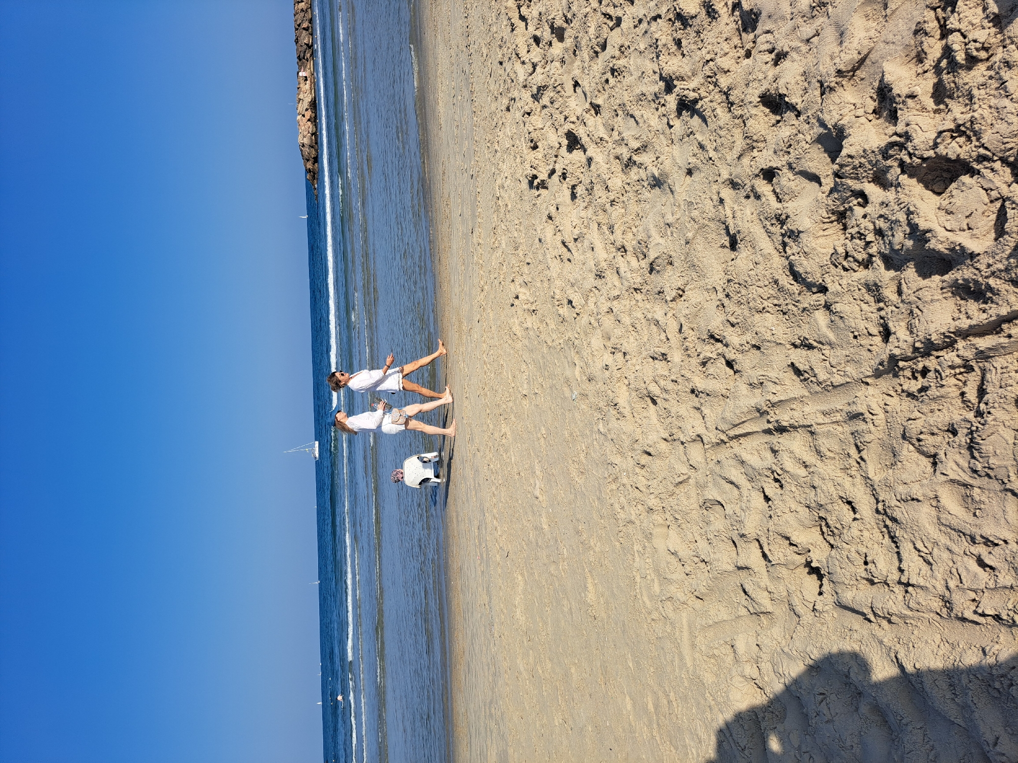 Two women are walking along the beach in Herzliya, Israel. The woman on the left is wearing a white top and shorts, while the woman on the right is wearing a white top and denim shorts. Both women are wearing sunglasses. In the background, a small white dog is sitting in a white plastic chair in the shallow water. The sea is calm with small waves, and several sailboats are visible in the distance. A rock jetty extends into the water on the right side of the image. The sky is clear and blue. The sand in the foreground shows footprints. The shadow of a person taking the photo is visible in the bottom left corner.
