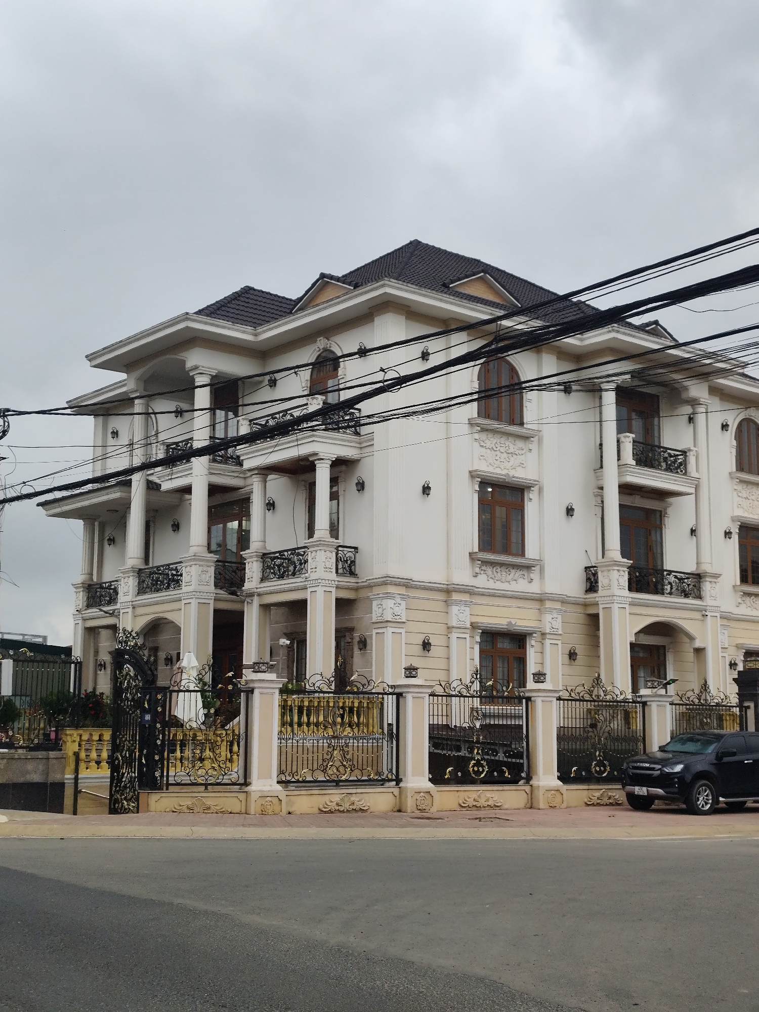 A large, opulent, multi-story house stands on a street in Cam Ly - Da Lat, Vietnam. The house is painted white with cream-colored decorative accents and features numerous classical columns, balconies with ornate black iron railings, and arched windows with brown frames. The roof is dark tile. A black SUV is parked in front of the house next to a decorative black and gold metal fence. The sky is overcast and grey, and a tangle of electrical wires runs across the top of the image.