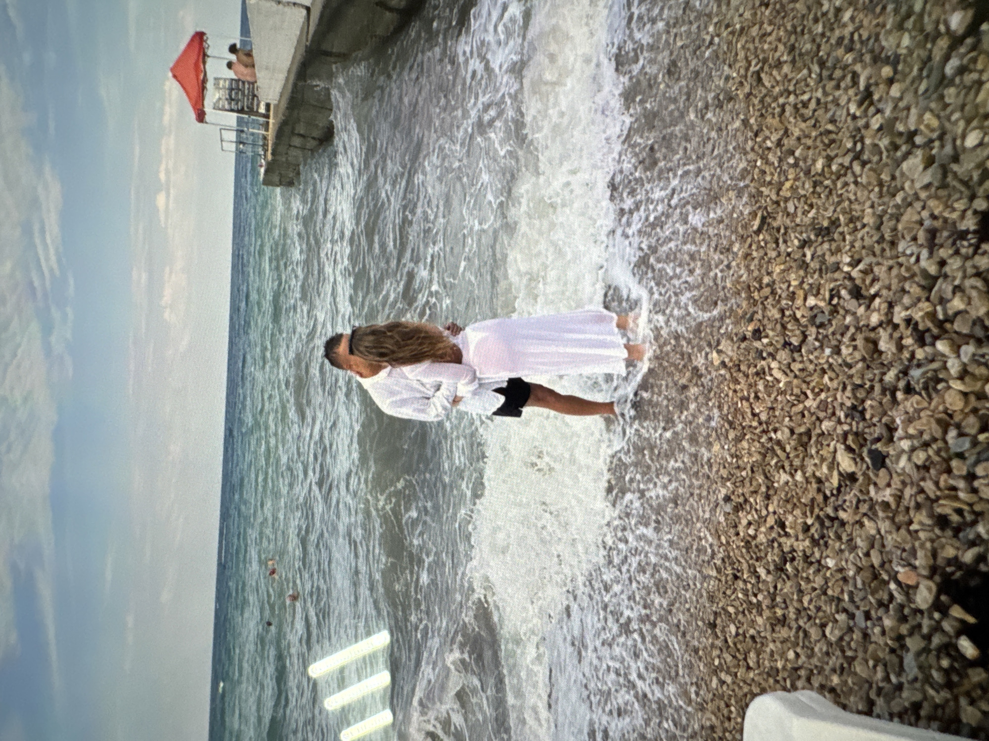 A couple is embracing on a pebble beach at the edge of the sea. The man is wearing a white shirt and black shorts, while the woman is wearing a long white dress. They are standing in the shallow water, with waves lapping around their feet. In the background, the sea is choppy under a cloudy sky. To the right, a concrete pier extends into the water, with a red umbrella and two people sitting beneath it. In the distance, several other people can be seen swimming in the sea.