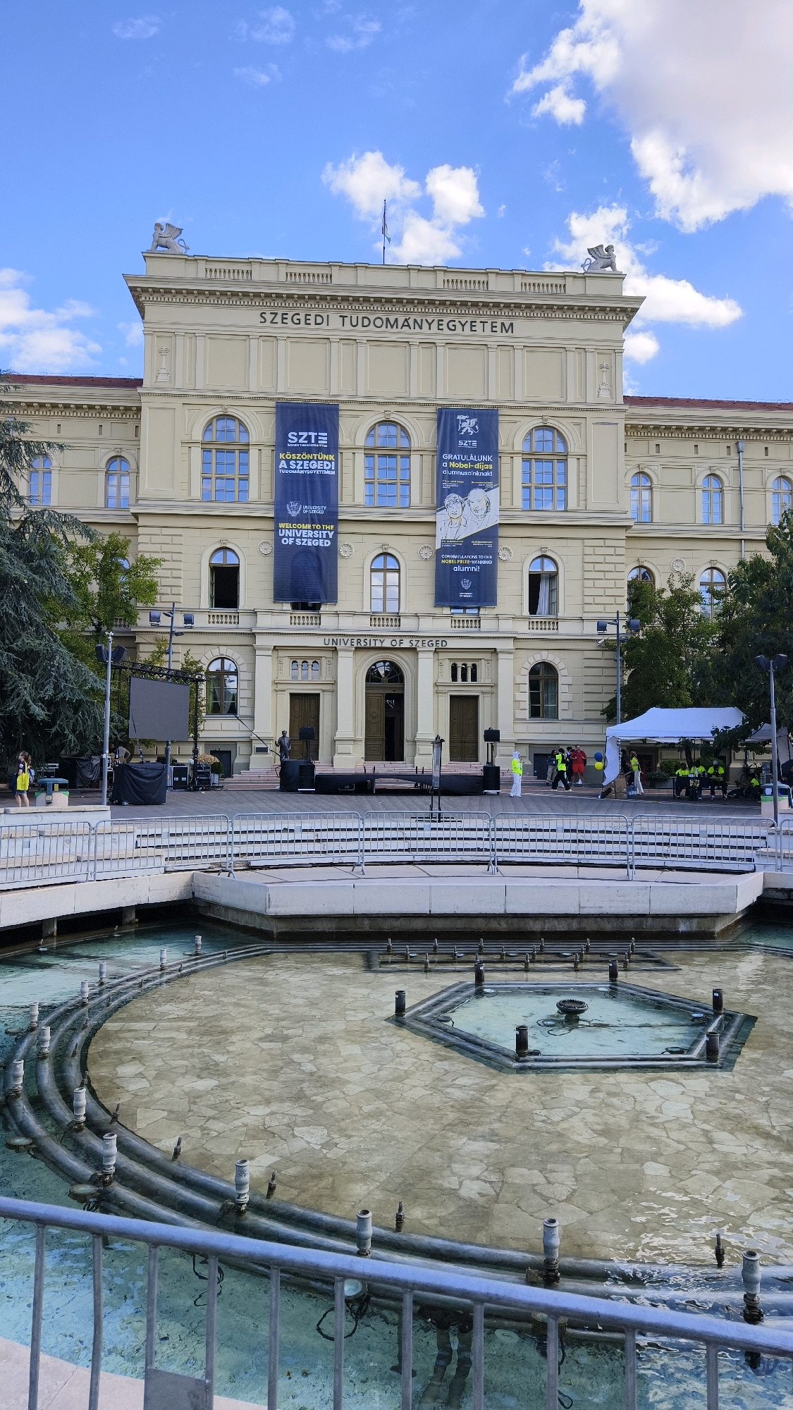 The image shows the front of the University of Szeged in Szeged, Hungary. The building is a large, light-colored neoclassical structure with multiple arched windows and a prominent inscription across the facade that reads "SZEGEDI TUDOMANYEGYETEM". Two large blue banners hang from the building, one displaying the text "KÖSZÖNTÜNK A SZEGEDI TUDOMÁNYEGYETEM", and the other "GRATULÁLUNK a Nobel-díjas", suggesting an event or celebration. Below these banners, more text reads "WELCOME TO THE UNIVERSITY OF SZEGED" and "CONGRATULATIONS TO OUR WORLD-RENOWNED ALUMNI!".
In front of the building, there is a wide staircase leading up to the main entrance, flanked by large double doors. To the left of the entrance, a large black LED screen is set up on a stage, along with audio equipment. Several people are visible in the foreground and middle ground. On the left, a person wearing a yellow top walks away from the camera. Near the stage, several individuals in high-visibility vests are present, some appearing to be setting up or managing equipment under a white tent structure.
In the foreground, a drained fountain is visible. It has a geometric design with multiple pipes and nozzles. A metal railing separates the fountain area from the viewer. The sky above is blue with scattered white clouds. The overall scene suggests an outdoor event or preparation for one at the university.