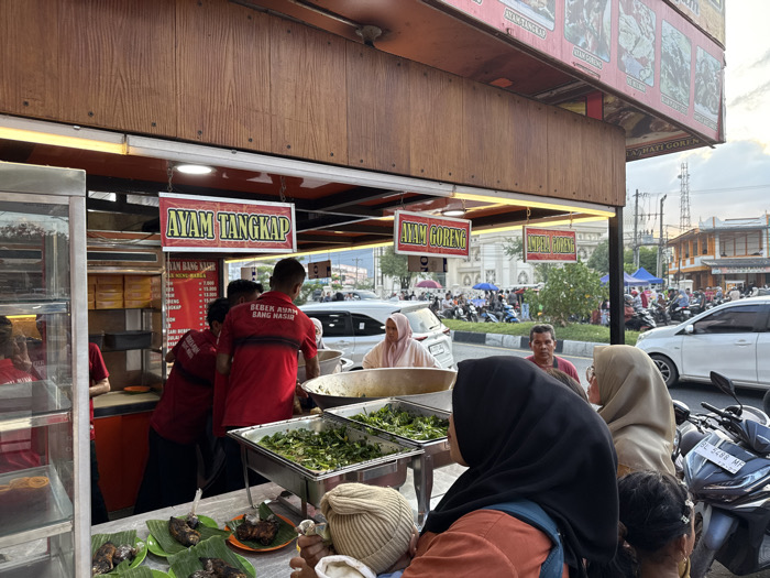 The image depicts a street food stall, identified as "BEBEK AYAM BANG NASIR," operating at dusk or early evening in Lambaro, Indonesia. The stall features a wooden facade with internal illumination and red-bordered signs displaying menu items such as "AYAM TANGKAP," "AYAM GORENG," and "INDELA GORENG," along with a visible price list.
Behind the counter, at least three male employees wearing red shirts with the stall's logo are engaged in preparing and serving food. Large metal trays containing green vegetables are prominently displayed, alongside individual portions of grilled fish or chicken served on small plates with banana leaf garnishes. A glass display case on the far left contains other food items.
Multiple customers are queued at the counter. In the immediate foreground, a woman wearing a black hijab and an orange top carries a baby in a front-mounted carrier. Next to her, another woman in a light beige hijab is visible. A man in a red t-shirt stands behind these customers, partially facing the viewer. Another woman in a light pink hijab is visible further to the left.
The background reveals a busy urban environment. A wide multi-lane street with a median strip is occupied by cars and numerous motorcycles, indicating active traffic and parked vehicles. In the mid-distance, a large white building with prominent arched architectural features stands. Further down the street, additional buildings and several temporary market stalls with colored awnings are visible, suggesting a vibrant market area. The sky is overcast, typical of evening conditions. Power lines and utility poles are also present.