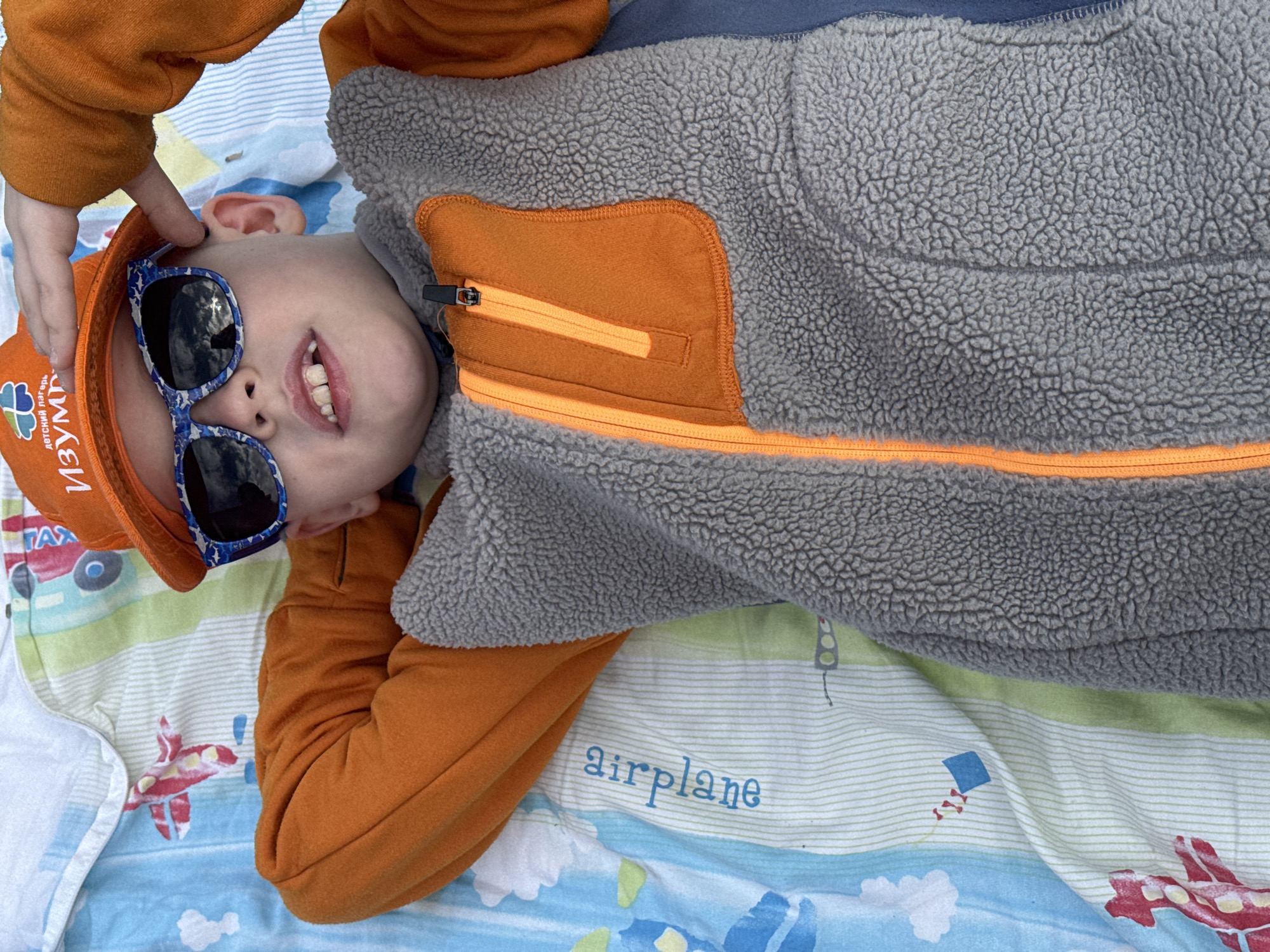 A young child lies on a patterned blanket, wearing an orange hat, star-patterned sunglasses, and a gray fleece jacket with orange accents. The hat bears the inscription "ДЕТСКИЙ ЛАГЕРЬ ИЗУМРУД" (Children's Camp Emerald) in Russian. The blanket features images of airplanes, cars, and the word "airplane." Based on the hat inscription, the location may be Chaikovsky, Russia, as it refers to the "Emerald" children's camp. The child is smiling and appears to be touching their hat.