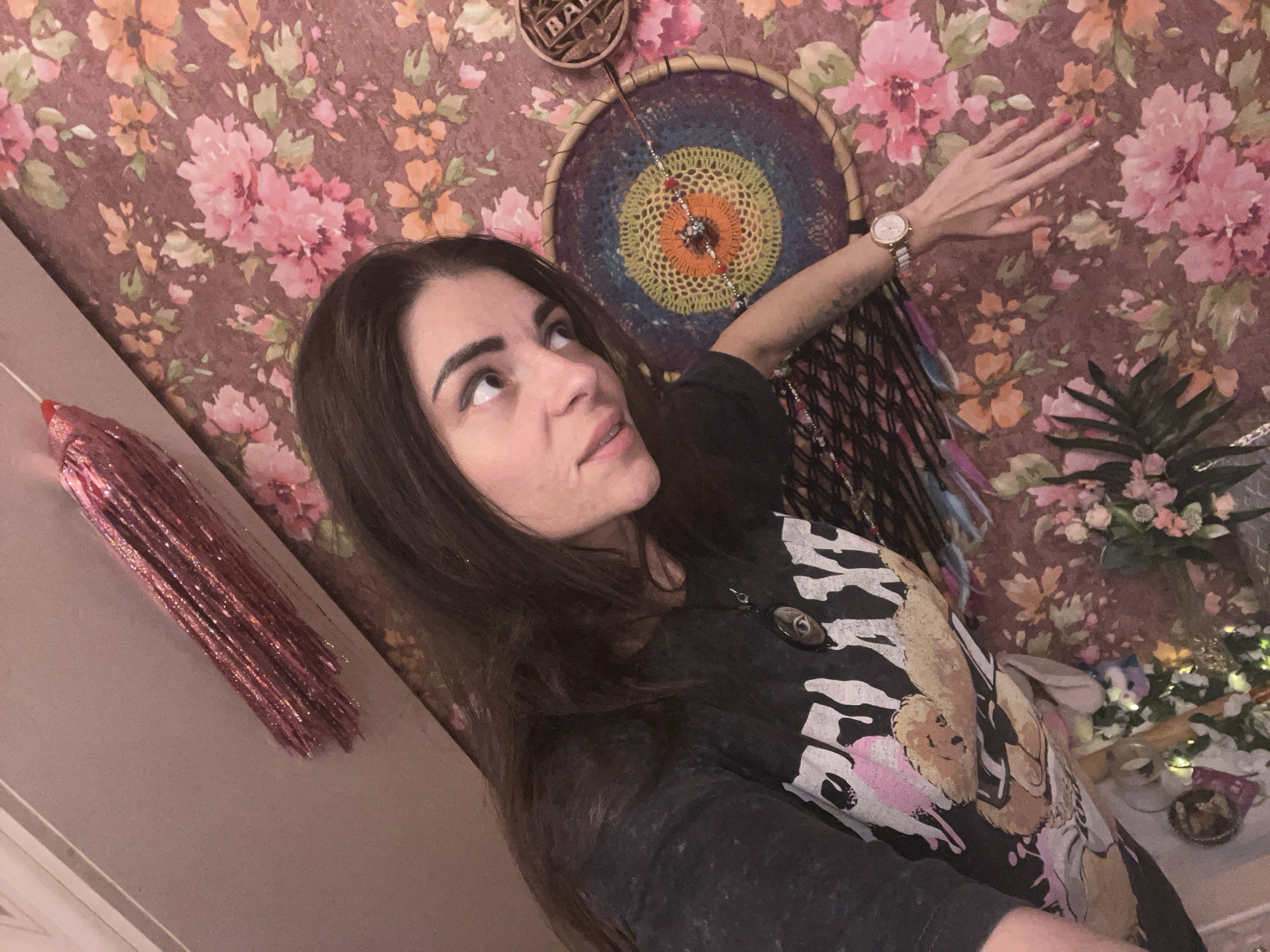 A woman with long, dark hair is shown in a selfie, looking upwards and to her left. She is wearing a dark t-shirt with a graphic print. Her right arm is extended, and she is wearing a wristwatch. Behind her, the wall is covered in floral wallpaper with pink and orange flowers. A dreamcatcher with colorful threads hangs on the wall to her right. To the left of the woman, a pink, glittery fringe decoration is visible against a neutral-colored wall. The lighting is warm and appears to be indoor. The setting does not provide any specific clues to its location in Krasnoe-na-Volge, Russia.