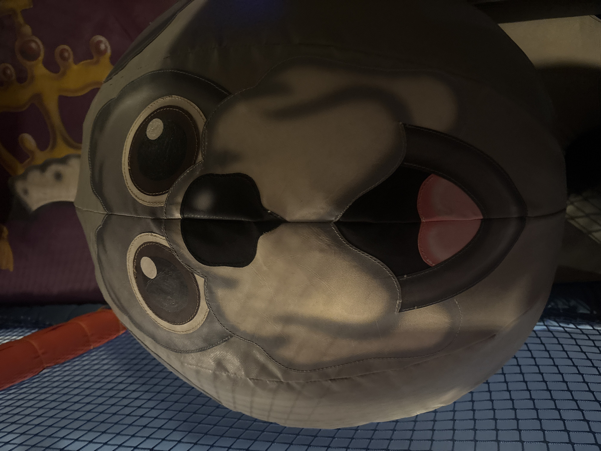 A close-up, low-angle shot shows a large, round, plush toy that resembles a cartoonish dog's head. The toy is gray with large, dark brown eyes with white pupils. Its nose is black and bulbous, and its mouth is open, revealing a pink tongue. The toy is situated in an indoor play area, possibly a soft play center. To the left of the toy is a blue net enclosure, and above it, a red inflatable tube. In the background, there are colorful decorations on a purple wall, including yellow, blob-like shapes. The lighting appears to be artificial.