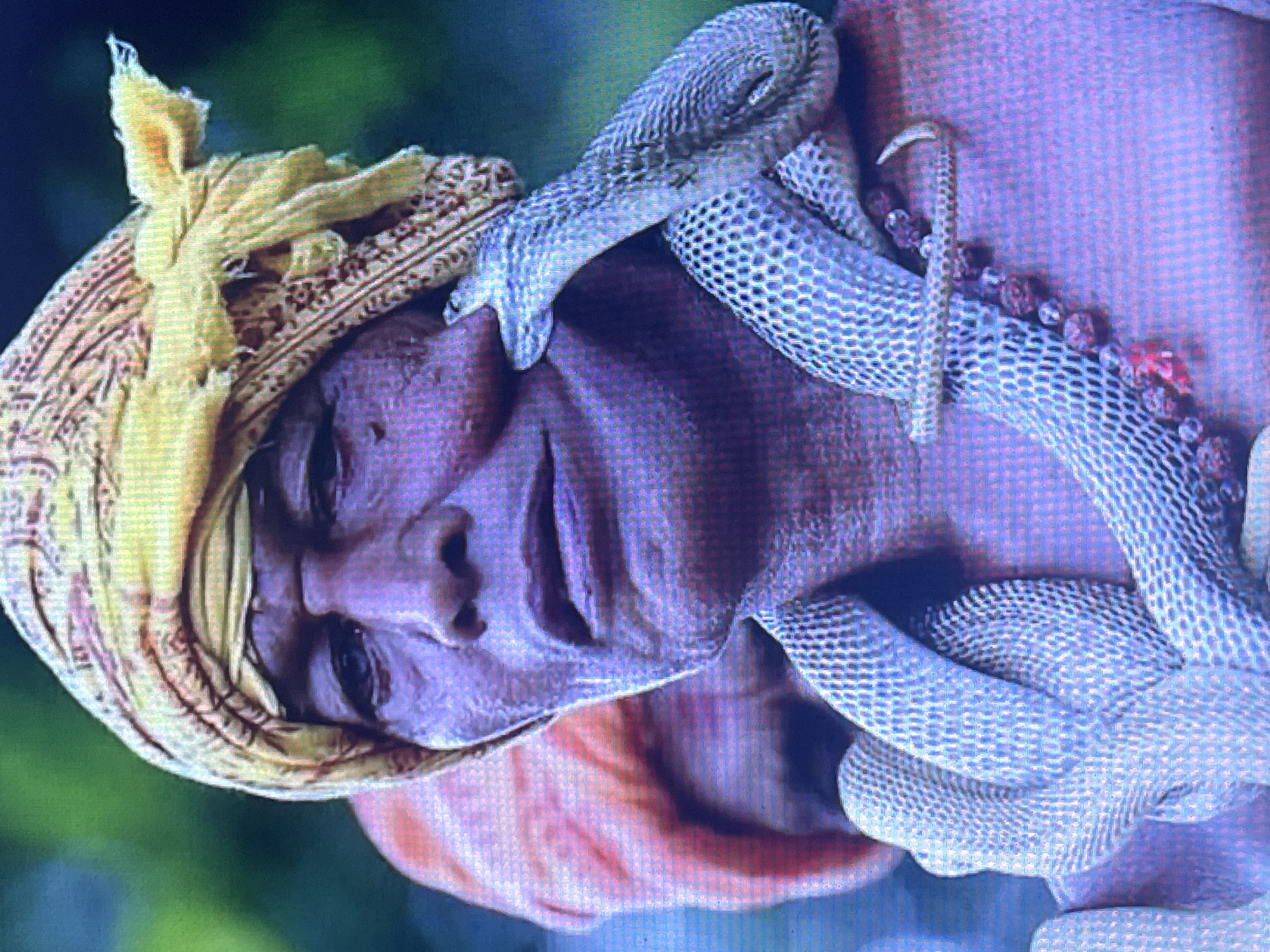 The image depicts a close-up of a man wearing a yellow turban and a necklace of beads. A light-colored snake is coiled around his neck and is touching his face. Another person wearing an orange turban is visible in the background. The environment appears to be outdoors with blurred greenery in the background. The man's facial expression is neutral. The lighting is diffused, casting a purple hue over the man's skin. The image's resolution is low, and it appears to be a photograph. There is no indication of the city of Stary Oskol, Russia, or any specific urban environment.