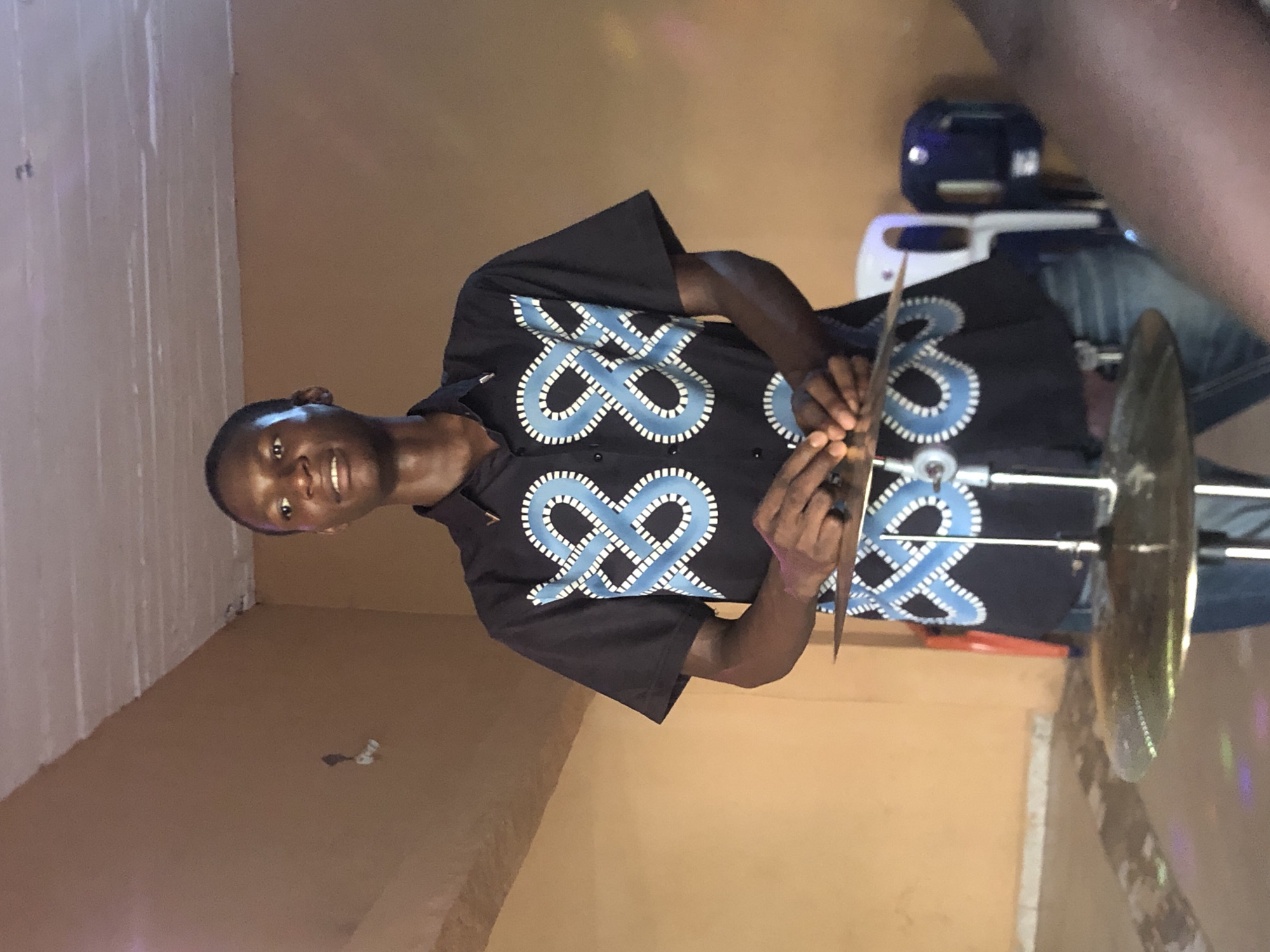 A man stands indoors in Nkwelle Ezunaka, Nigeria. He wears a black shirt patterned with blue and white designs and is holding a pair of cymbals. There is a stand holding another cymbal, and a blue object is visible in the background. The walls are painted in shades of brown and white.