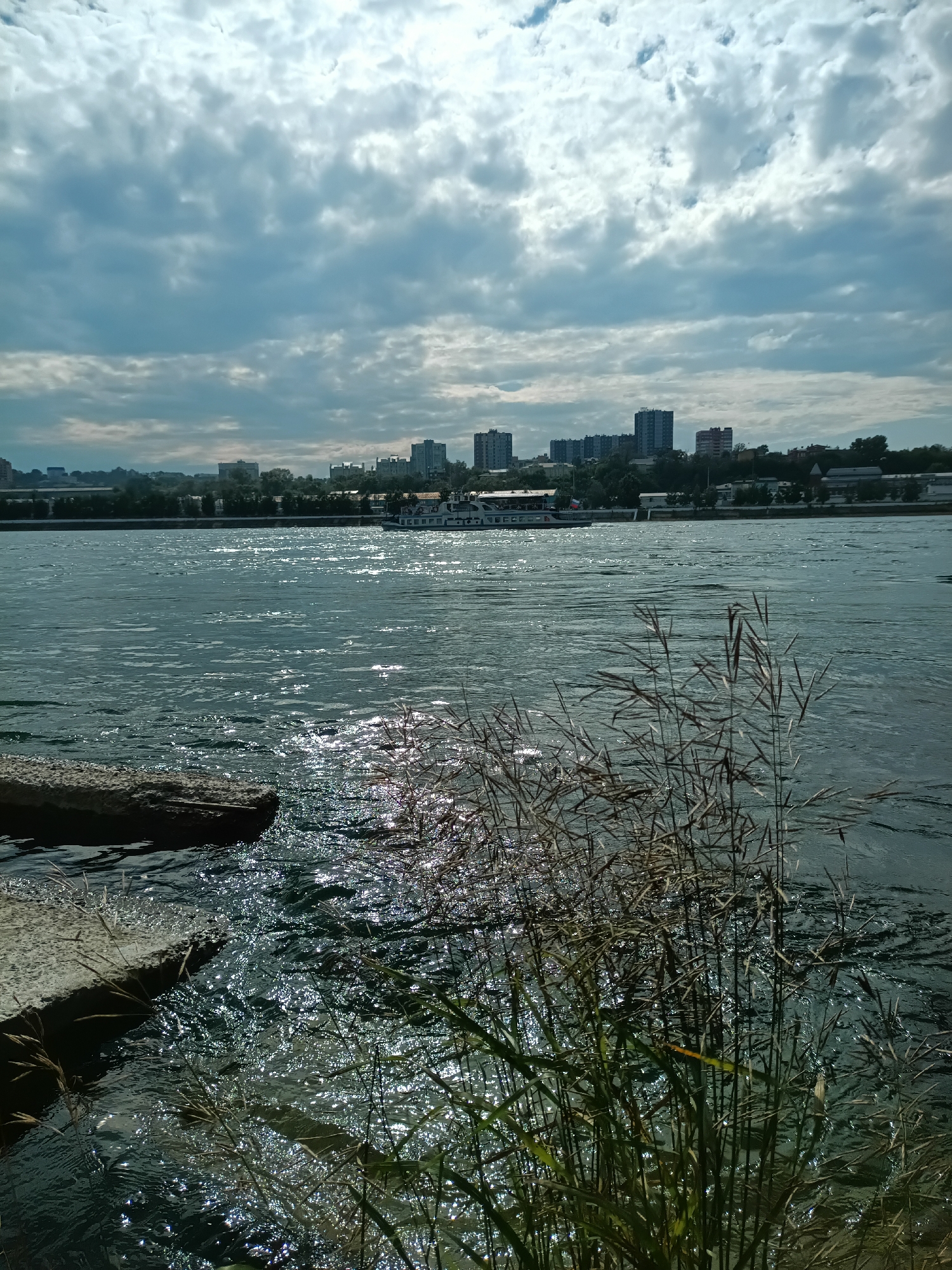 The image depicts a wide, choppy river in Irkutsk, Russia, under a cloudy sky. The foreground features concrete elements, possibly a bank or pier, and tall, dry grasses swaying in the breeze. In the middle ground, a white passenger boat with a red flag is visible, traversing the water. The background showcases the city skyline, characterized by a cluster of tall, modern buildings and trees lining the riverbank. The sunlight reflects off the water's surface, creating a shimmering effect. No people are visible in the frame.