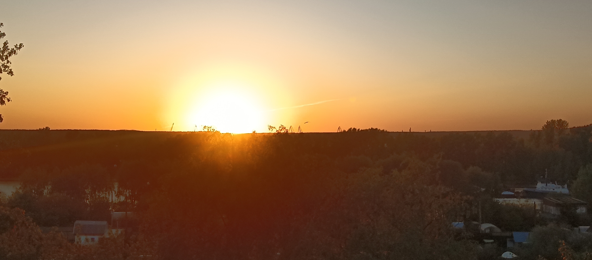 A wide-angle shot depicts a sunset or sunrise illuminating a landscape in an unknown city. The sun, a radiant orb, sits low on the horizon, casting a bright yellow-orange glow across the sky. The horizon line is marked by a dark silhouette of land, with what appears to be structures, possibly cranes or transmission towers, punctuating the skyline. Below the horizon, a dense layer of trees and foliage dominates the midground, obscuring much of the ground below. Scattered buildings and possibly a small body of water can be seen amongst the trees in the lower part of the frame. The light from the sun creates a strong contrast between the brightly lit sky and the darkened landscape below.