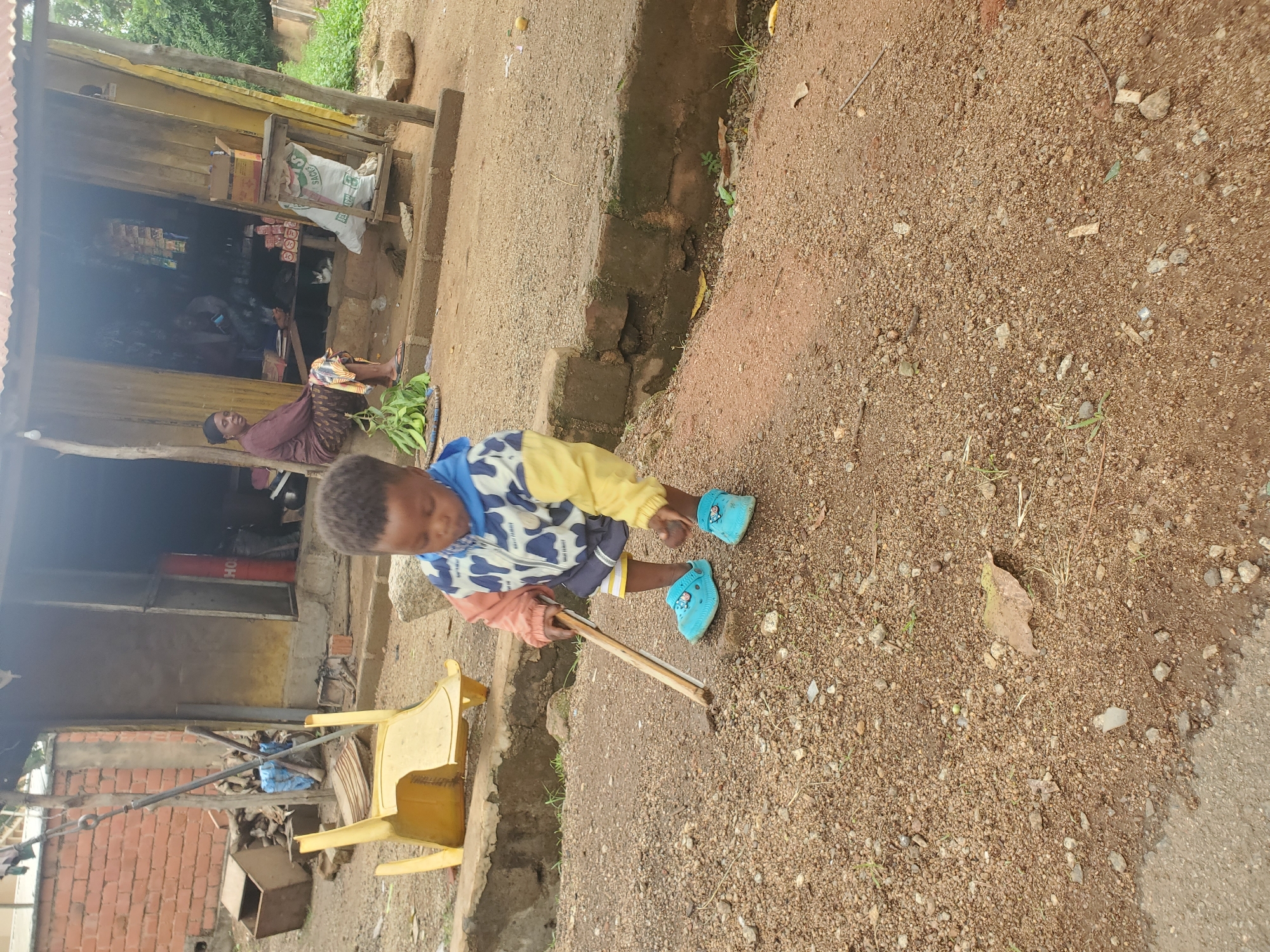 A young child, wearing a colorful jacket and blue Crocs, is standing on a gravelly path in front of a small shop. The child is holding a wooden stick, appearing to dig or play with the ground. Behind the child, a yellow plastic chair is overturned. The shop, constructed from wood and metal sheeting, has a few items displayed outside. Inside the shop, a woman is seated and another person is partially visible. The scene is outdoors, with a brick wall on the left and vegetation visible in the background. The environment suggests a rural or semi-urban setting.