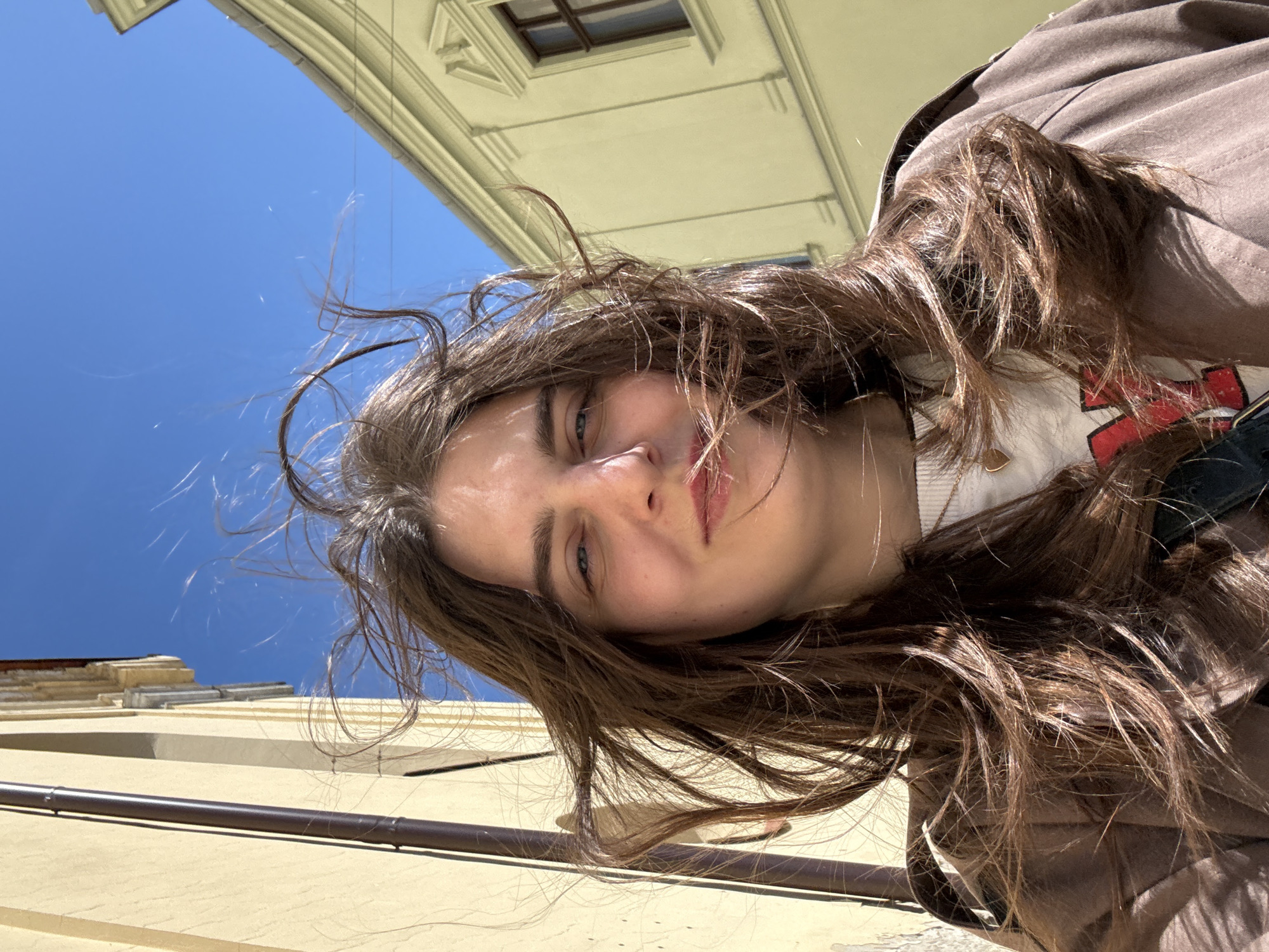 A low-angle selfie featuring a young woman with long, wavy, light brown hair partially obscuring her face. She wears a light-colored sweater and a brown jacket. Behind her, buildings with light yellow facades and a blue sky are visible, suggesting an outdoor setting. The image is oriented at an unusual angle. Based on architectural details, the location is likely Trnava, Slovakia.