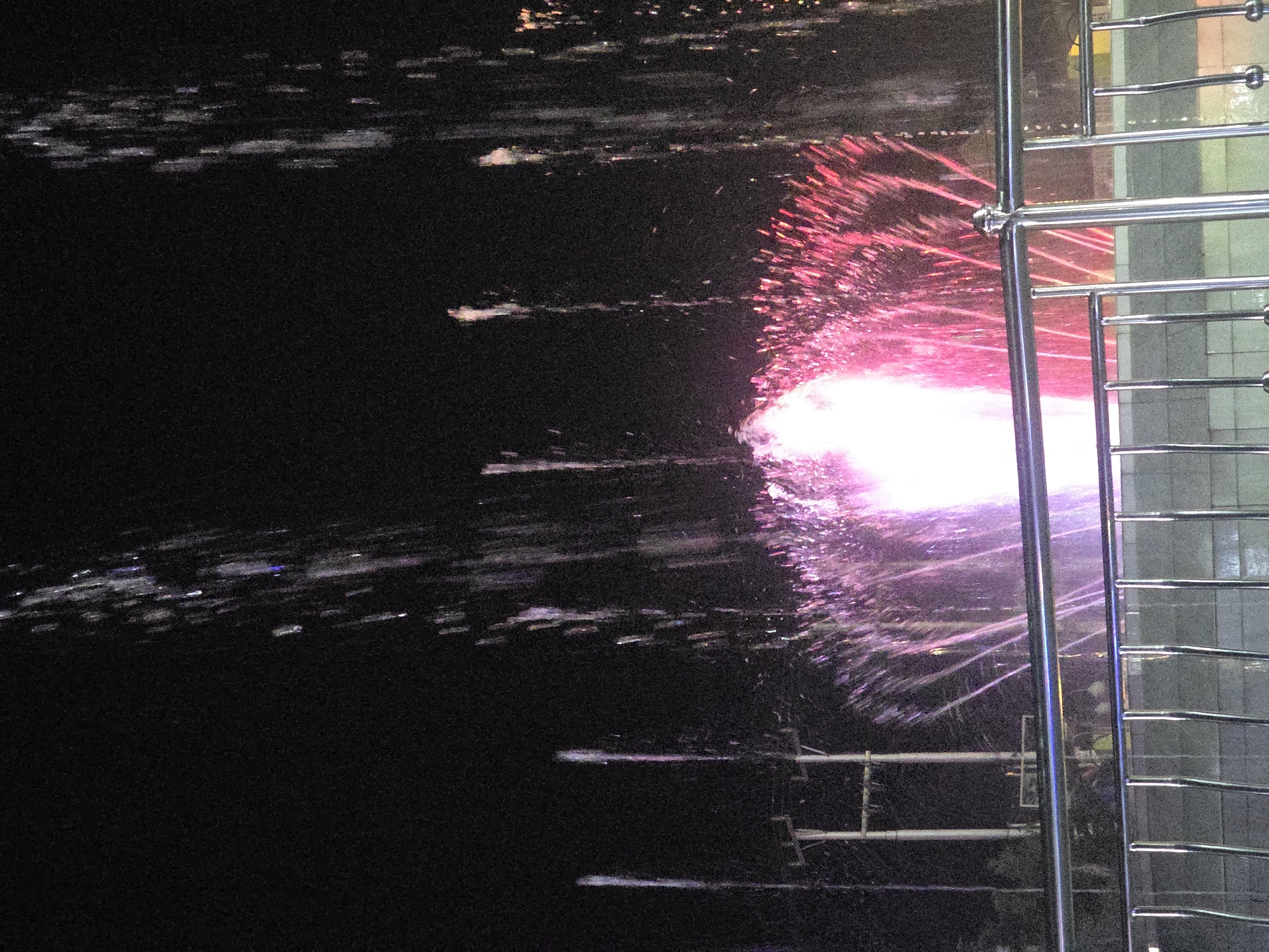 The image depicts a nighttime scene of a fountain in the city of Padang Aro, Indonesia. The fountain is illuminated with bright pink and white lights, creating a vibrant spray of water against the dark sky. Several vertical streams of water are visible, some illuminated with white and purple hues, while others are a darker, less defined mass. In the background, there are utility poles and wires, suggesting an urban setting. A polished metal railing runs across the foreground, with vertical supports and horizontal bars. Through the railing, the base of the fountain structure and some indistinct figures are visible, possibly people watching the display. The overall impression is one of a public space at night, with the fountain as the central attraction.