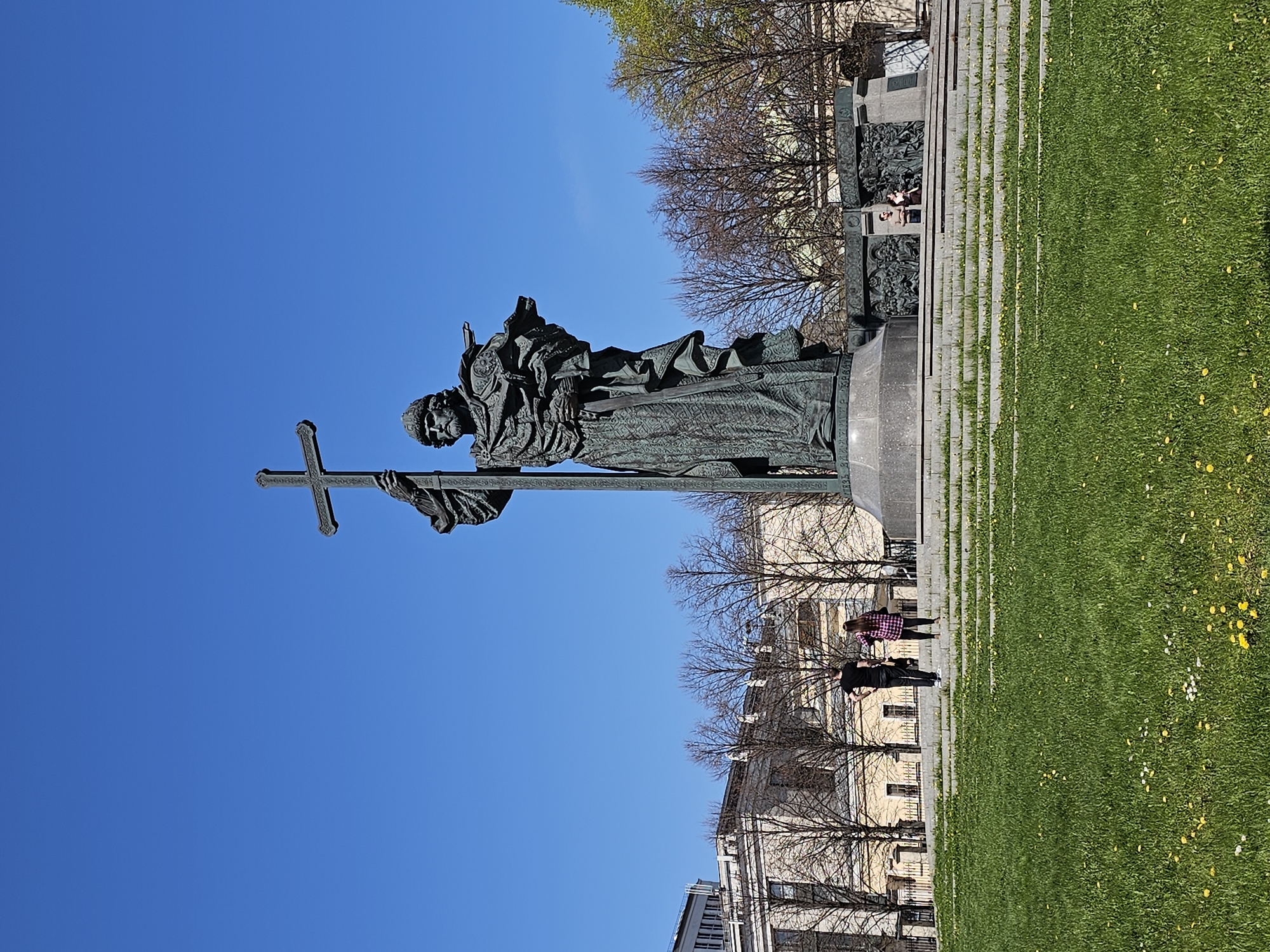 The image depicts a bronze monument of a religious figure holding a cross, with a clear blue sky in the background. The statue is positioned on a stone plinth, with a stone staircase leading up to the monument. Patches of green grass are visible on the right side of the image, dotted with small yellow and white flowers.
Several bare trees are interspersed between the statue and the buildings in the background. Some individuals are standing on the lower portion of the steps and along a pathway, suggesting the statue is in a public square or park setting. The architecture of the buildings in the background suggests an urban environment.
Based on the statue's design and the overall context, it is highly likely that this image shows the Monument to Saint Vladimir in Moscow, Russia, which was unveiled in November 2016. The monument is located near the Kremlin, on Borovitskaya Square.