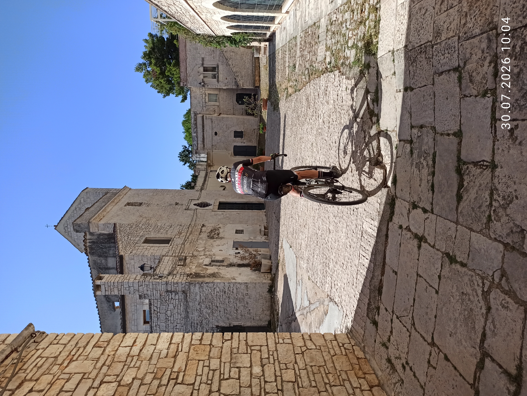 A person wearing a cycling outfit and helmet rides a bicycle through a cobblestone courtyard. The buildings in the courtyard are made of light-colored stone with rustic architectural details. The sky is clear and blue, and the sun casts shadows on the ground. The timestamp "30.07.2026 10:04" is visible in the lower right corner of the image. The scene appears to be in a historic town square in Terlizzi, Italy.