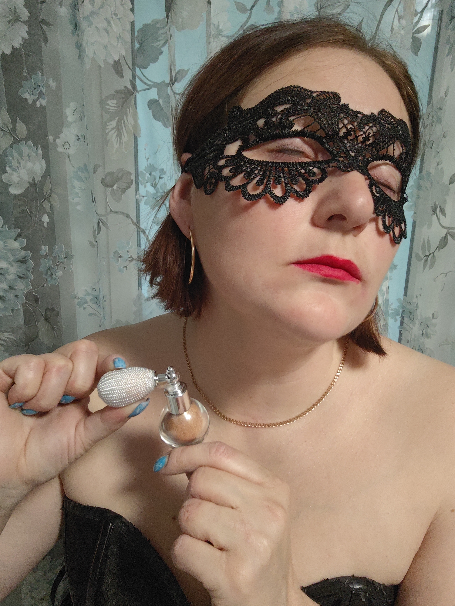 A woman wearing a black lace mask is holding a perfume atomizer with a silver bulb. She has red lipstick and gold jewelry (earrings and necklace). Her nails are painted blue with a white pattern. She is wearing a strapless black garment. The background features a floral patterned curtain. The location is not specified, but it is not Markovskii, Russia.
